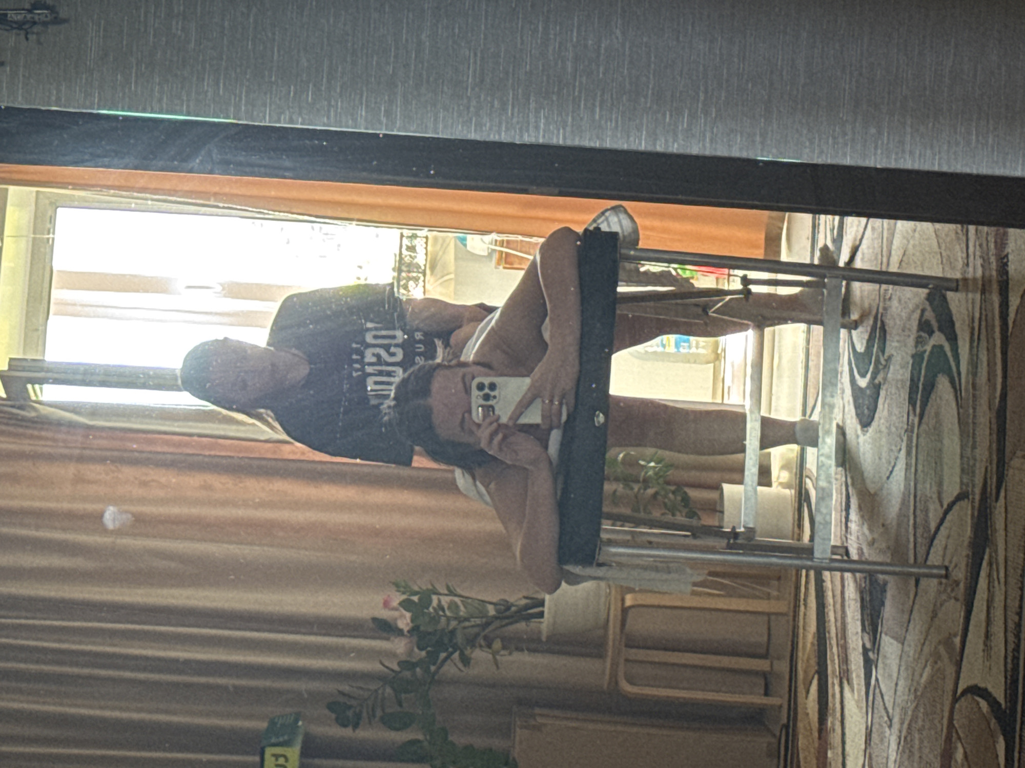 A photograph depicts the reflection of a couple in a mirror. The woman is holding a smartphone up, presumably taking the picture. She is wearing a light-colored top. The man is standing behind her, wearing a dark t-shirt with some writing visible on it.
The reflection also shows a portion of the room they are in. A clothing rack is positioned next to the couple. Behind the rack is a floral arrangement, and in the background, one can see parts of the room, including a window and some furniture. A patterned rug is on the floor.
The location is Chaikovsky, Russia.