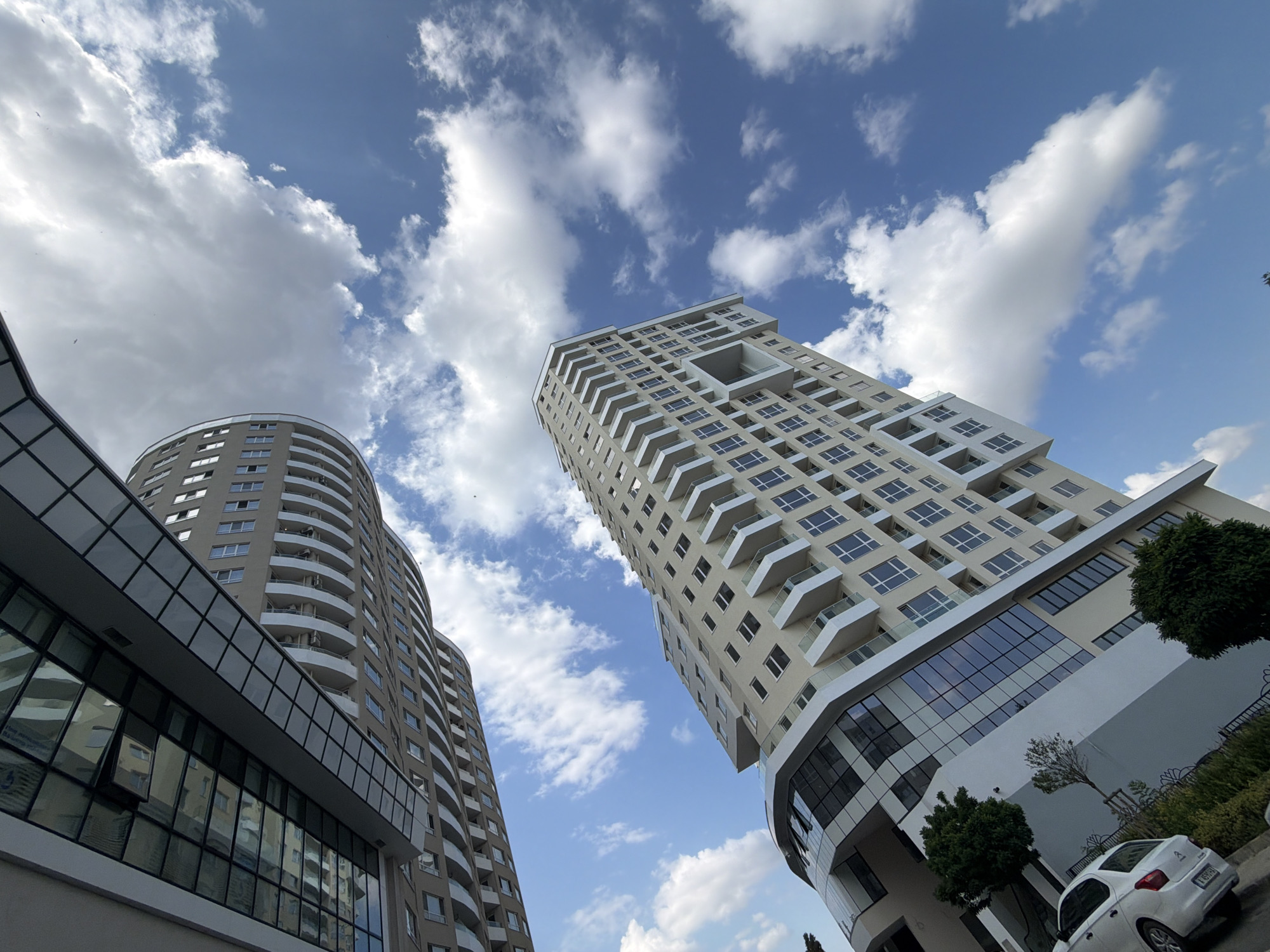 The image depicts two modern apartment buildings in Burgas, Bulgaria, set against a bright blue sky with scattered white clouds. The building in the foreground is a tall, slender structure with light-colored facade, numerous windows, and protruding balconies with white railings. The lower section of this building features a curved glass facade. To the left and behind, another, wider building is partially visible. This building has a darker facade and a prominent glass-covered walkway or connecting structure. The perspective is from a low angle, looking upwards at the buildings. In the lower left corner, a white car is parked, and some green foliage is visible. The overall scene suggests a contemporary urban environment in Burgas.
