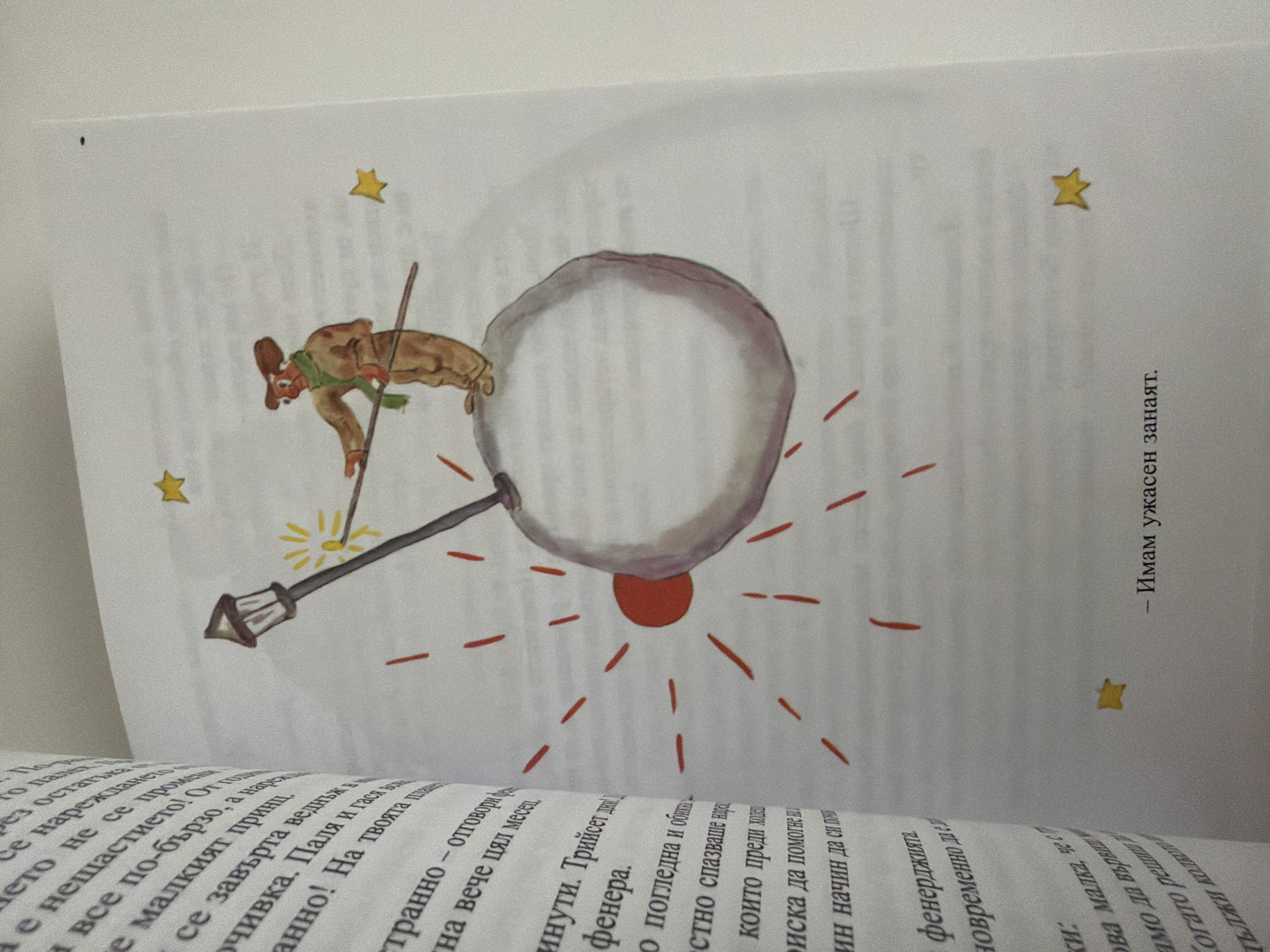 The image displays an open book with text on the left page and an illustration on the right page. The illustration depicts a small, barren planet with a single lamppost. A figure in a brown outfit and green scarf stands on the planet, holding a stick that appears to be lighting the lamppost. There are red lines radiating from a red circle on the planet, suggesting light or an explosion, and a few yellow stars are scattered on the white background. The text on the left page is in Bulgarian. The image does not contain any elements that would place it in the city of Pazardzhik, Bulgaria.