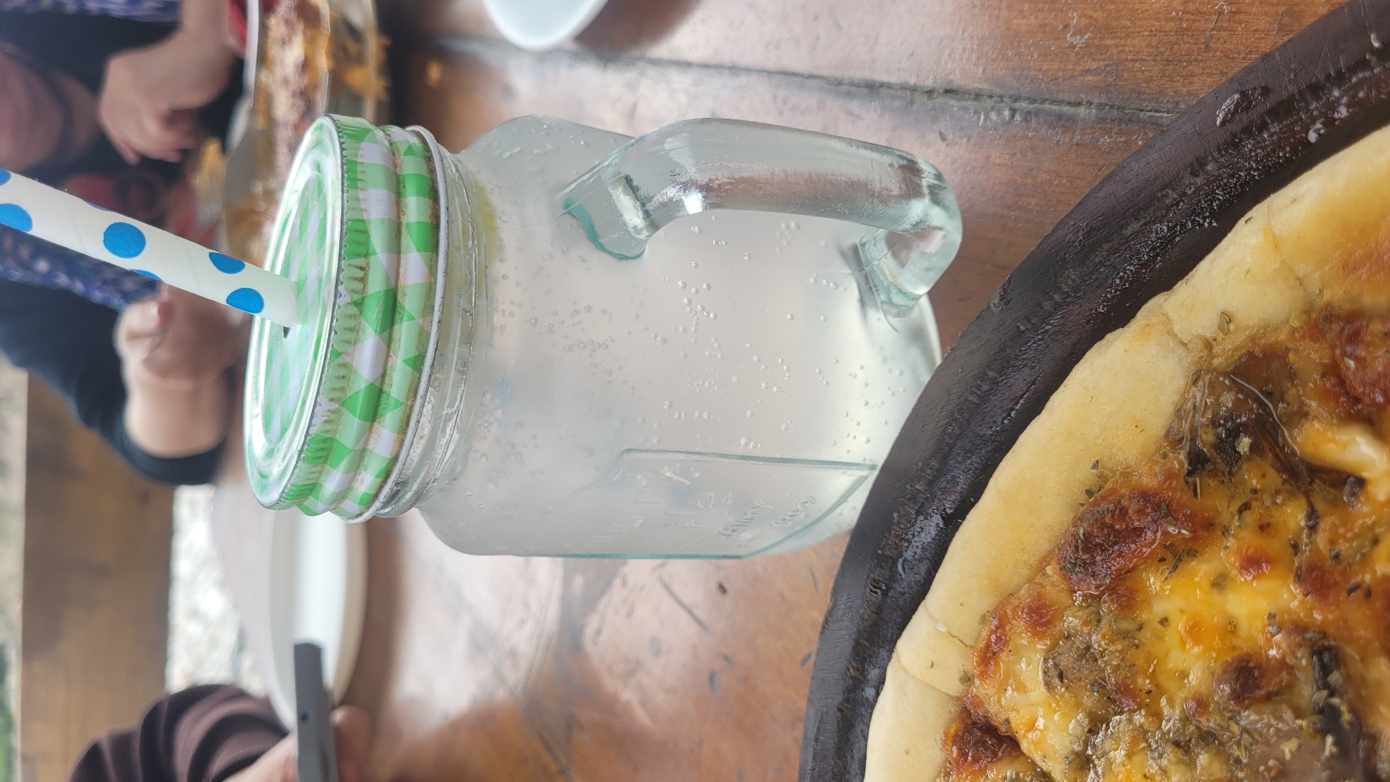 A close-up, low-angle shot focuses on a glass mason jar filled with a clear, bubbly beverage and a blue and white polka-dot straw. The jar has a green and white checkered lid and a handle. In the foreground, a slice of pizza sits on a dark wooden serving board, with visible cheese, herbs, and mushrooms. The background shows blurred figures of people at a table, suggesting a dining setting. The scene appears to be outdoors, with natural light. Based on typical imagery associated with such settings and known locations, this scene could plausibly be in Vashisht, India, a popular tourist destination known for its hot springs and guesthouses that often have open-air dining areas.