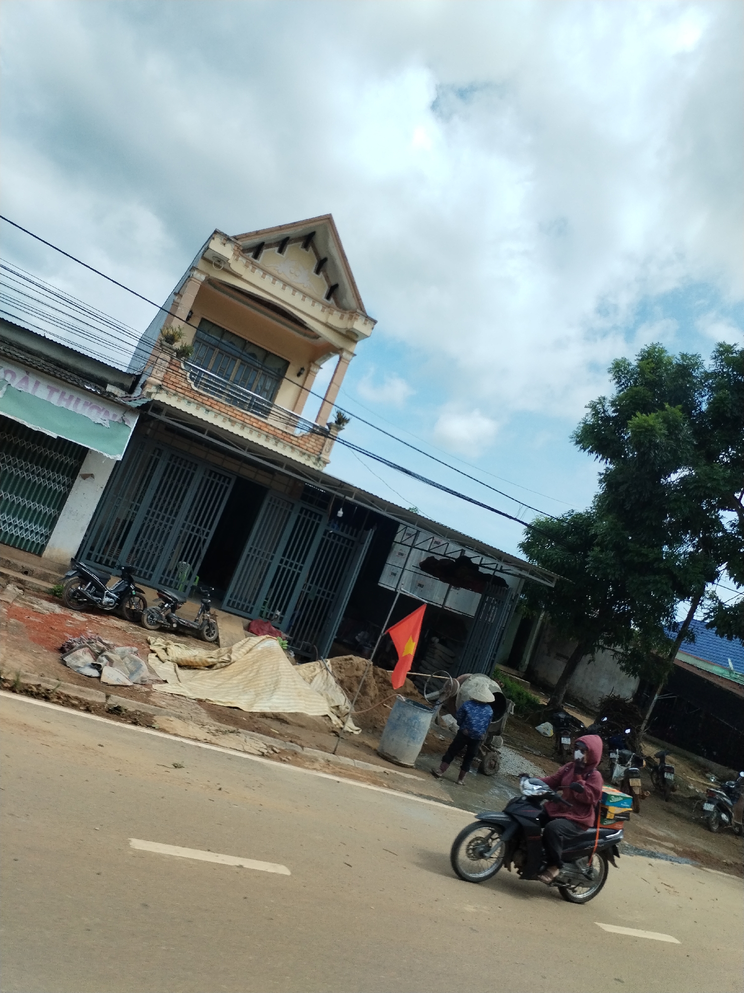 The image shows a street scene in Pờ Y, Vietnam. A two-story building with a faded yellow facade is angled in the frame. The ground floor features large, closed metal doors, and several motorbikes are parked nearby. Construction materials and equipment are scattered on the adjacent sidewalk, including a Vietnamese flag.
A person is operating a concrete mixer behind a blue barrel. Another person is on a black motorbike, driving along the road. A tree is in the background. The sky is partly cloudy.