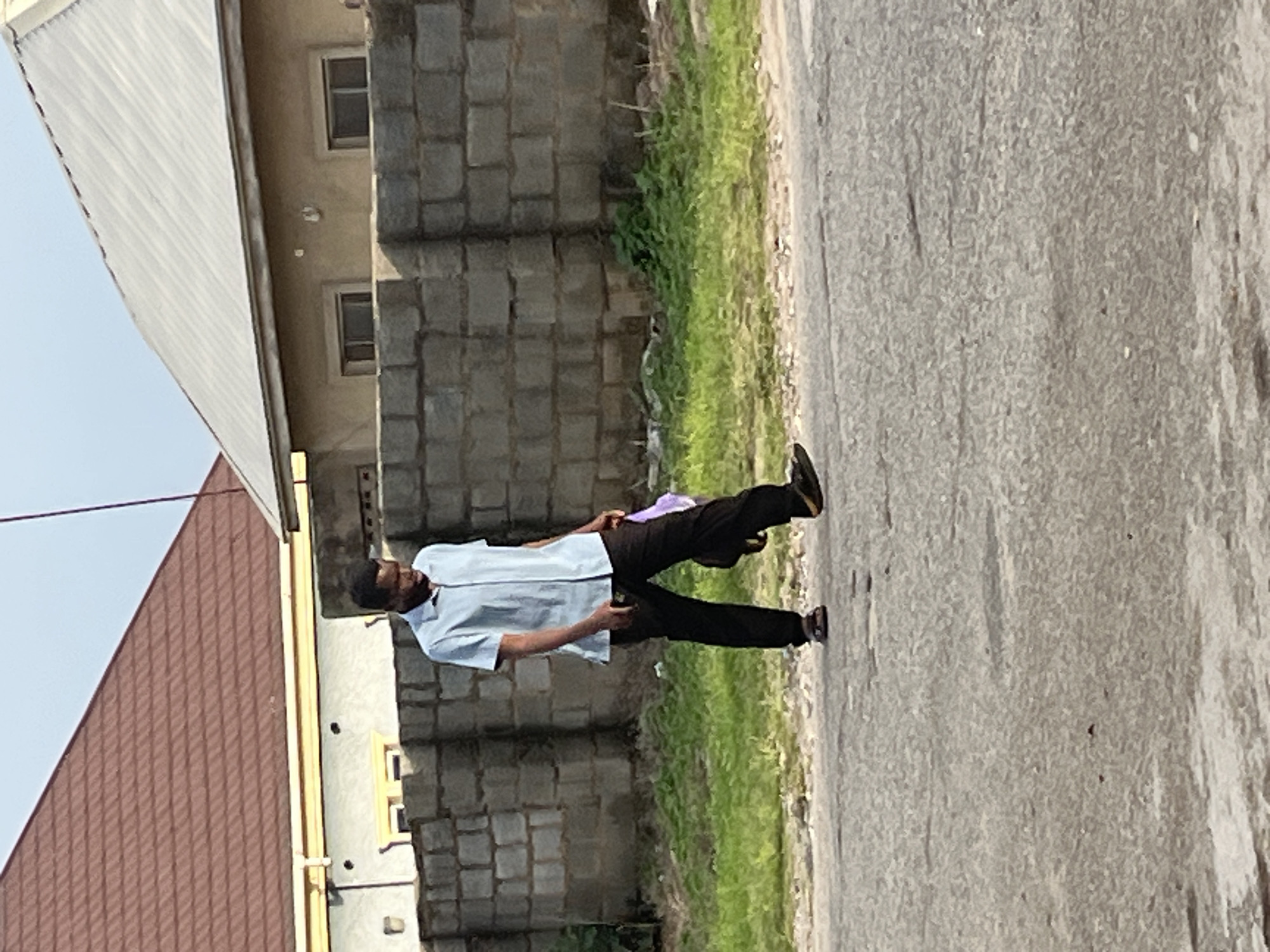 A young Black man walks on a paved road with a light blue short-sleeved button-down shirt and black pants. He carries a dark bag slung over his left shoulder and holds a purple item in his right hand. The man is wearing sandals. Behind him, a gray cinder block wall separates the road from an area of green grass and vegetation. Beyond the wall, two buildings are visible. The building on the left has a reddish-brown tiled roof and a light yellow facade. The building on the right has a silver corrugated metal roof and a light beige facade. The sky is overcast and pale blue. The scene appears to be in a residential area of Abuja, Nigeria.