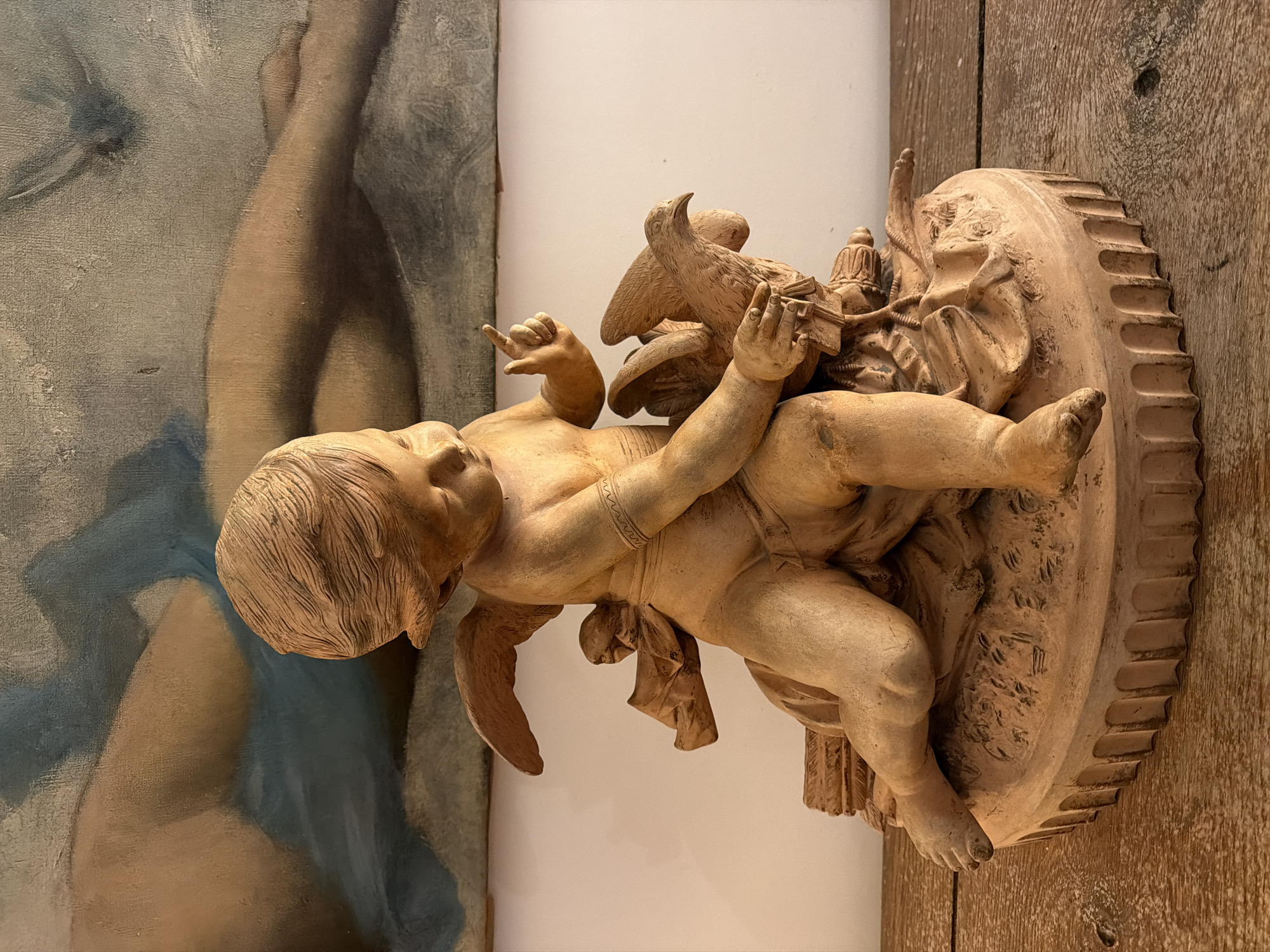 A terracotta-colored cherub statue, facing left with wings and perched on an ornate, circular base, is centered in the frame against a white wall. The statue is positioned in front of a section of a painting to the left and a weathered wooden surface to the right. The painting shows a person's torso and arm, while the wooden surface has visible wood grain and nail holes. The statue appears to be indoors. Without additional information, it is impossible to determine the location in Paris, France.