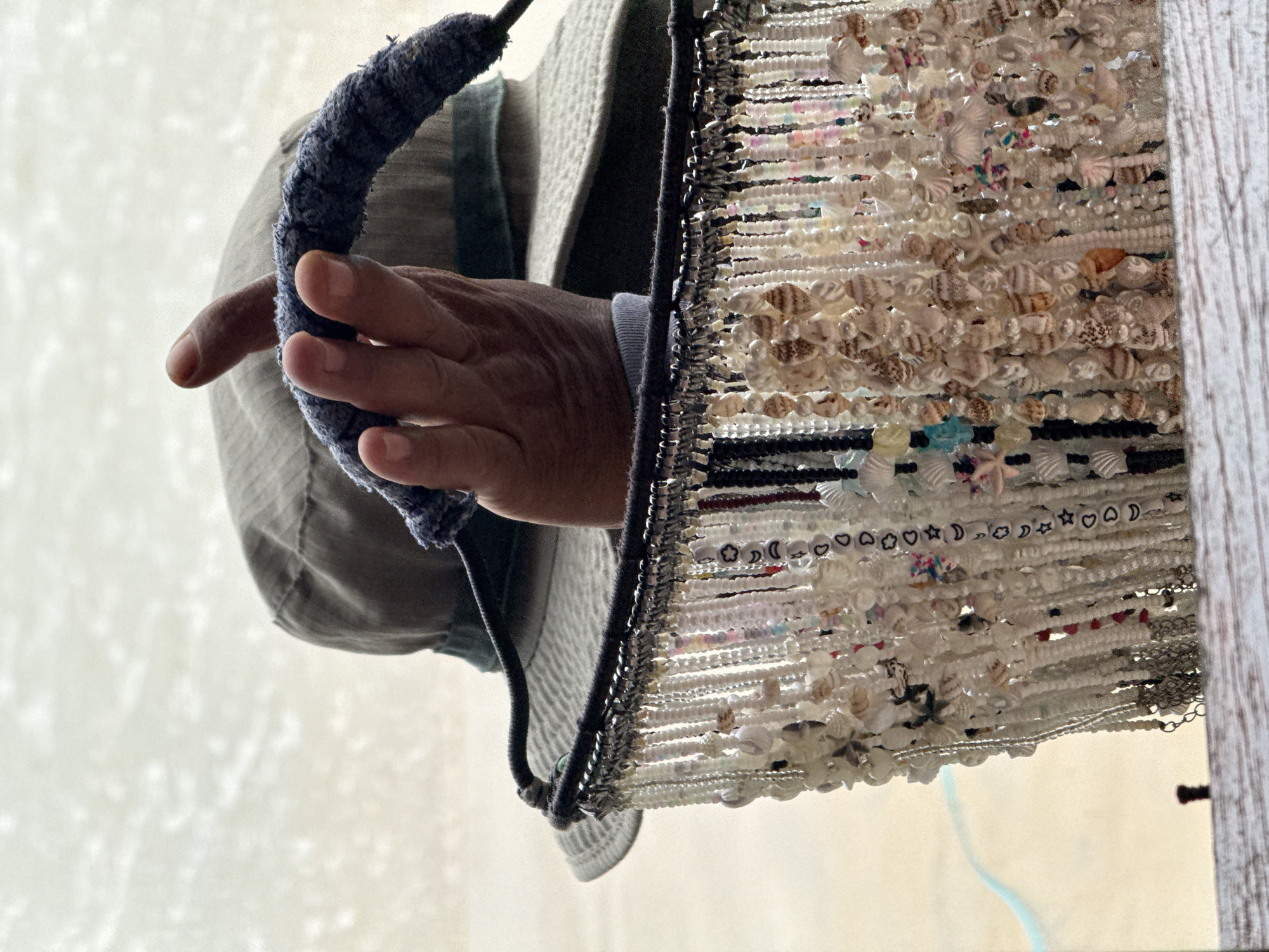 The image shows a close-up of a person's hands holding a collection of shell necklaces. The person is wearing a khaki hat and a light-colored shirt. The necklaces are made of small shells and beads, and they hang in multiple strands. The background is out of focus, but it appears to be a beach or a sandy area, possibly in Karon, Thailand. The lighting is bright, suggesting it is daytime.