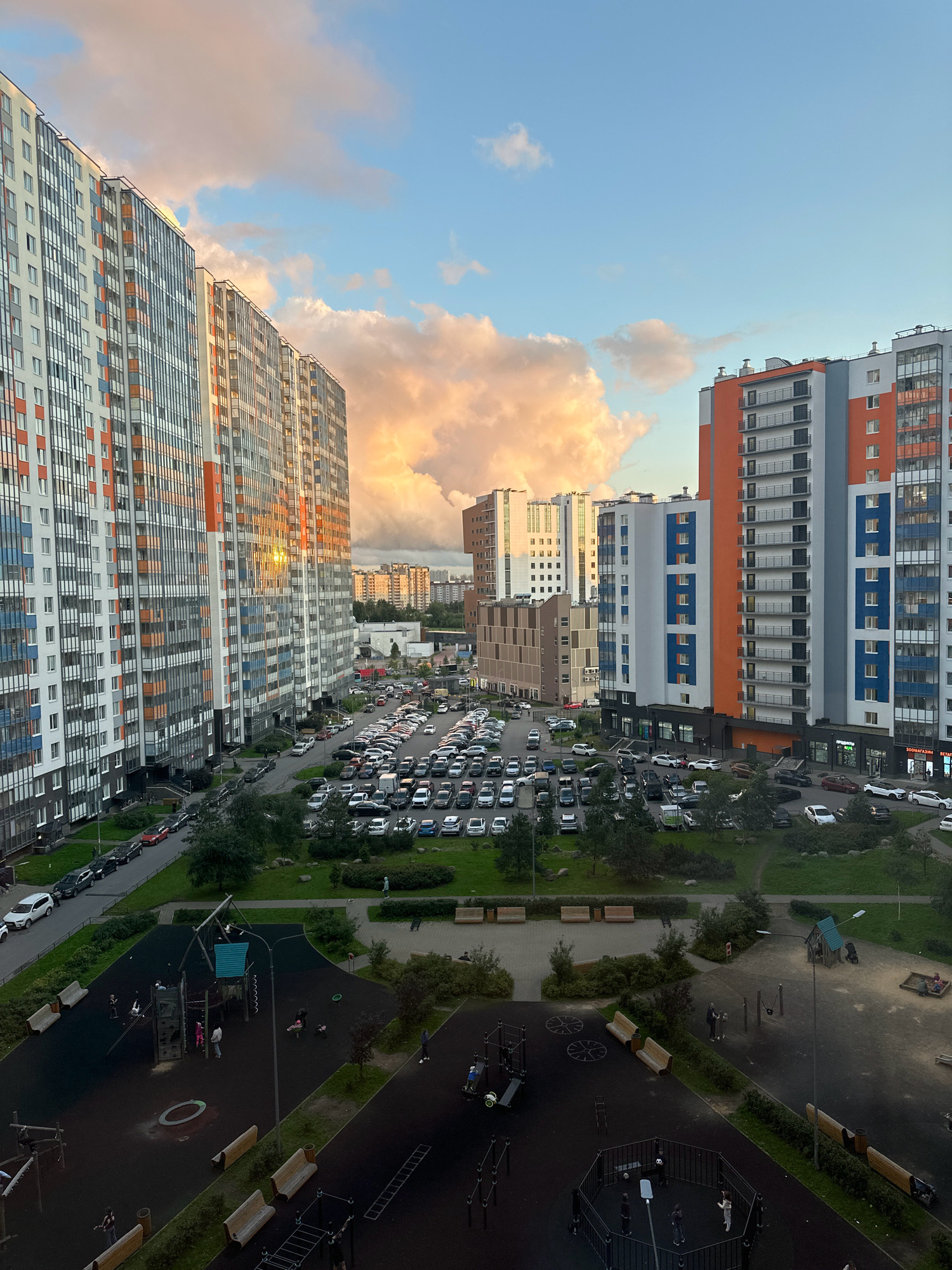 The image captures a view from a high-rise apartment building in Saint Petersburg, Russia, looking out over a residential courtyard. The sky is a mixture of blue with scattered clouds, some of which are illuminated with a warm, orange glow, suggesting it is either sunrise or sunset.
In the foreground, to the left and right, are portions of modern, multi-story apartment buildings with colorful facades, featuring balconies and numerous windows. The buildings on the left are predominantly white with vertical stripes of orange and grey, while the building on the far right has sections of orange, grey, and blue.
Dominating the mid-ground is a large, paved parking lot filled with a variety of cars. The cars are parked in rows, suggesting an organized parking system. Beyond the parking lot, more apartment buildings and some lower structures are visible, stretching into the distance.
The lower portion of the image shows a residential courtyard. There are children's playgrounds with various equipment, including swings, climbing structures, and a sandbox. Several benches are placed around the play areas and along pathways. Some individuals, including adults and children, are present in the courtyard, with some children playing and adults observing or walking. Green spaces with grass and trees are interspersed throughout the courtyard, offering a contrast to the built environment. The overall scene depicts a typical urban residential setting with elements of community spaces.