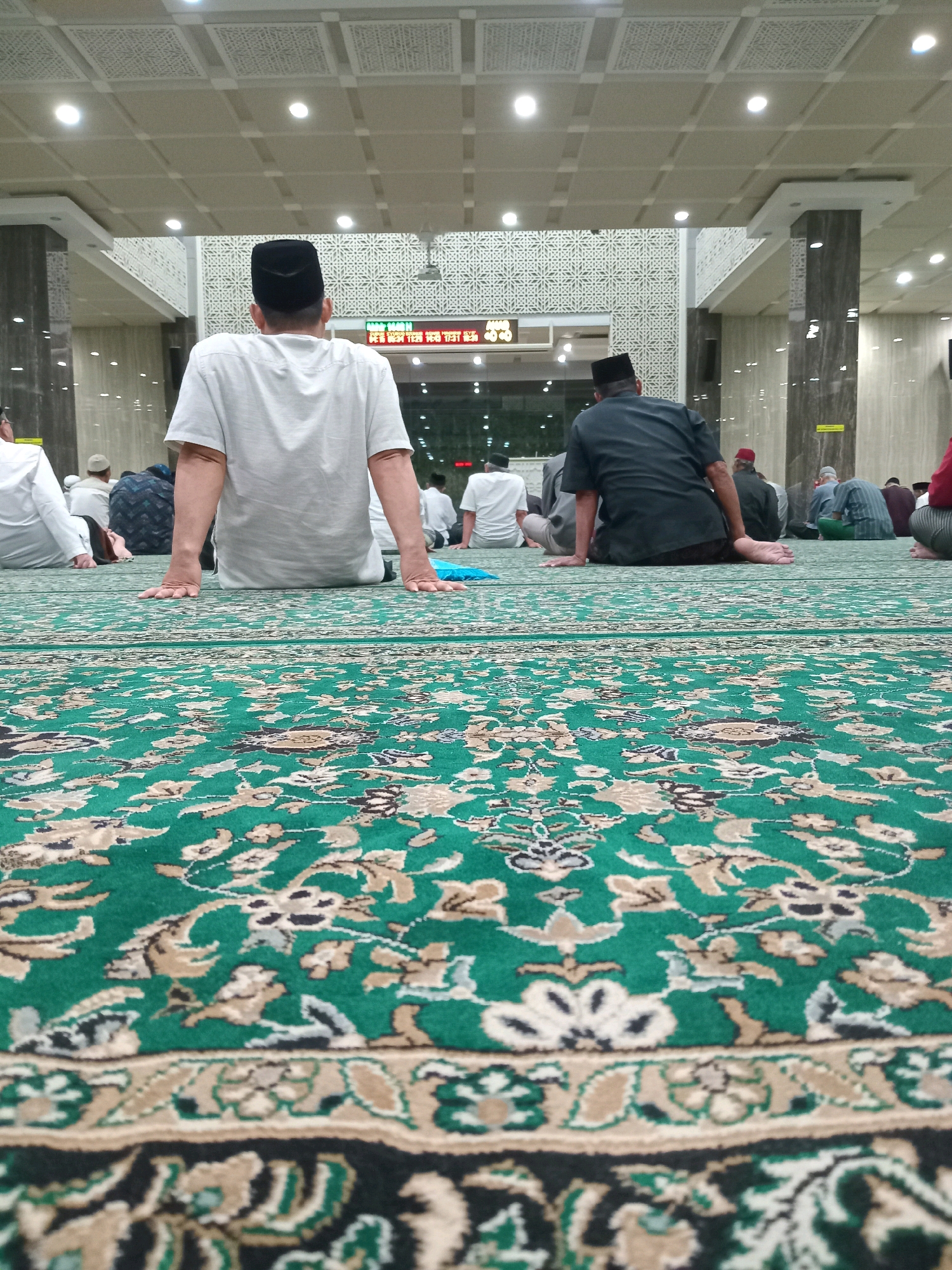 In Randuagung, Indonesia, a low-angle shot captures men sitting on a green carpet inside a mosque. The carpet features a floral pattern with cream, beige, and black accents. In the foreground, the hands of a person wearing a white shirt and a black skullcap rest on the carpet. Behind them, other men, also wearing skullcaps, are seated. The background reveals pillars, decorative walls with geometric patterns, and a ceiling with recessed lighting. A digital clock displaying numbers is visible in the distance. The scene suggests a religious gathering or prayer session within the mosque.