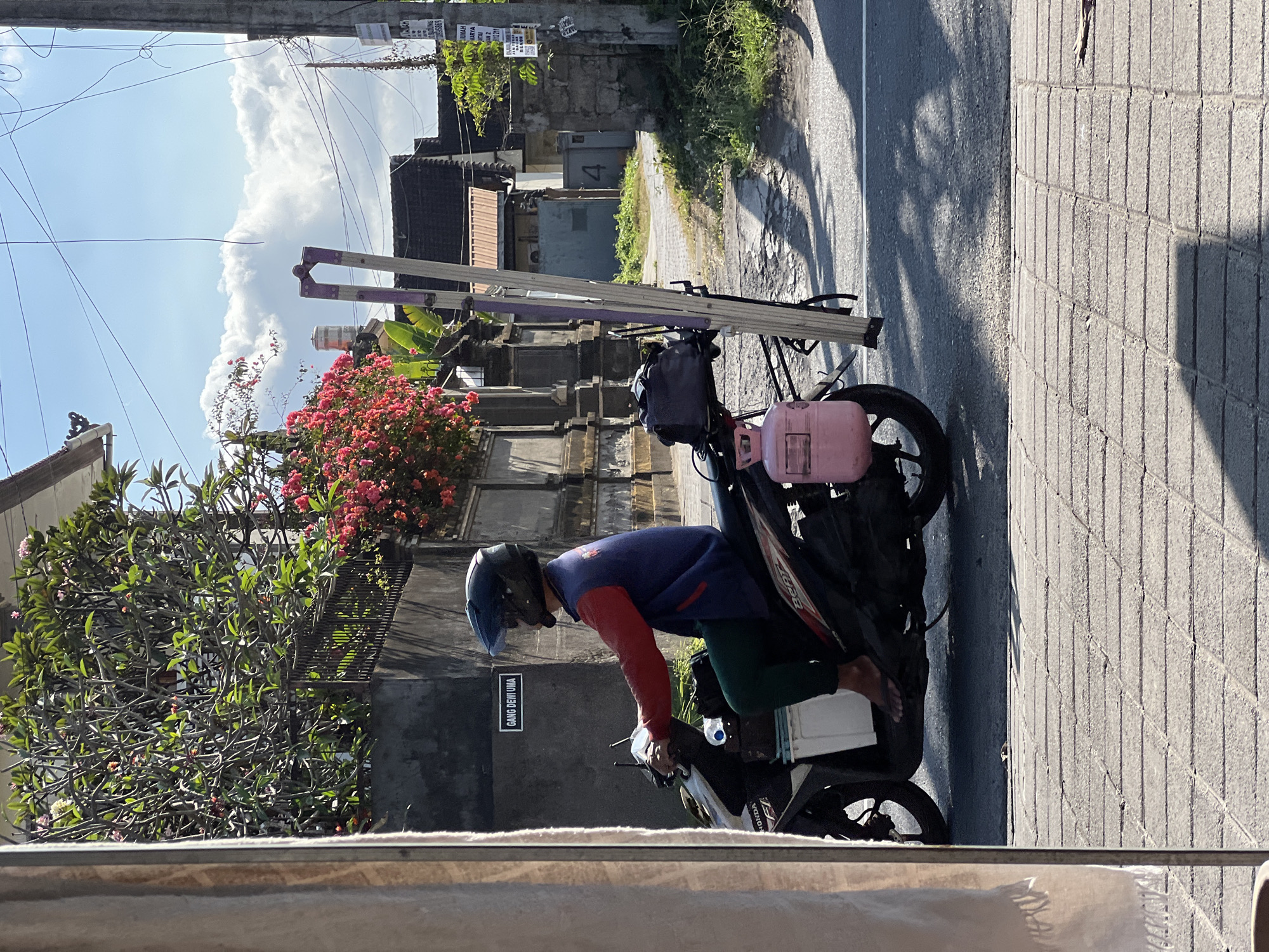 In Denpasar, Indonesia, a person wearing a blue vest, red shirt, and dark helmet rides a Honda Beat motorcycle. The motorcycle is equipped with a tall, foldable ladder strapped vertically to the rear, along with a pink propane tank and a black duffel bag. The rider is wearing green pants and flip-flops. The motorcycle is on a paved road adjacent to a sidewalk made of grey bricks. In the background, there is a wall with a sign that reads "GANG DEWI UMA," followed by lush green trees with pink flowers, and residential buildings with tiled roofs. The sky is blue with scattered white clouds. The scene suggests a typical day in a residential area of Denpasar, with the rider likely transporting equipment for a job.