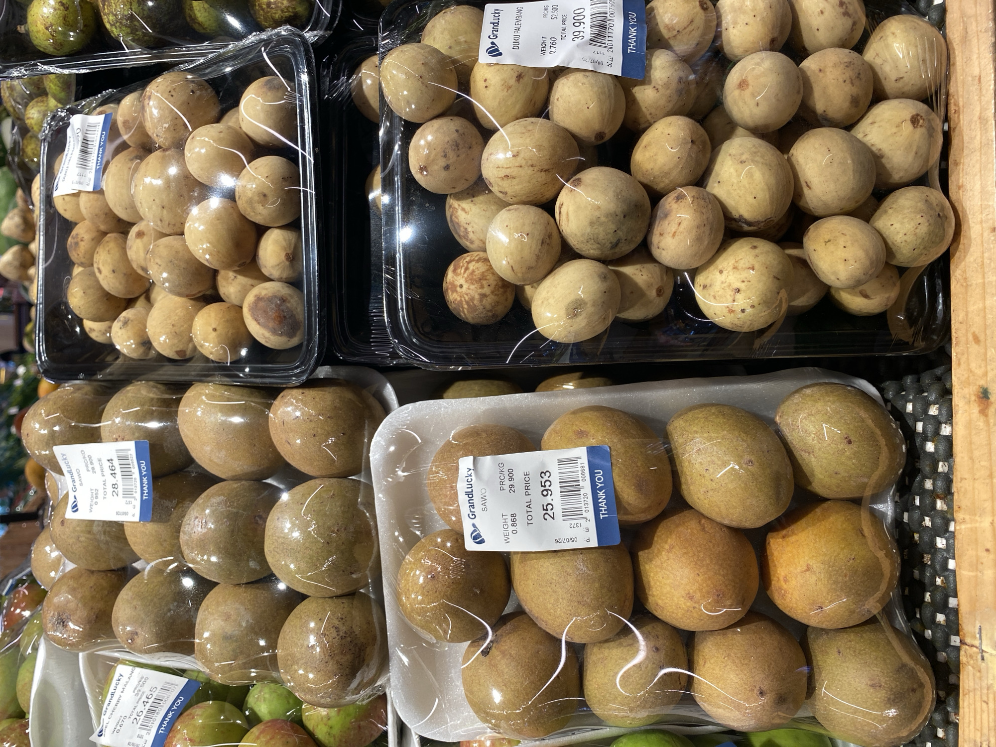 This image captures a close-up view of fruit for sale at a supermarket in East Jakarta, Indonesia. The focus is on several trays of tropical fruits, predominantly sapodilla and what appear to be langsat or duku.
In the foreground, on the left, a white styrofoam tray contains approximately ten sapodillas, each individually wrapped in clear plastic. A label on the tray indicates "GrandLucky SAWO" with details like "WEIGHT 0.868," "PRC/KG 29.900," and "TOTAL PRICE 25.953." Another label on the left, also with clear plastic-wrapped sapodillas, reads "GrandLucky SAPODILLA MALANG."
To the right, two black plastic trays are filled with smaller, round, light-yellow fruits, likely langsat or duku. These fruits are also individually wrapped in clear plastic. The label on the front tray reads "GrandLucky DUKU PALEMBANG" and shows "WEIGHT 0.760," "PRCKG 52.500," and "TOTAL PRICE 39.900." A similar tray is visible behind it, also containing the same type of fruit. In the background, more fruits are visible, including green and red fruits on the left, potentially apples or other produce.
The overall environment suggests an indoor retail setting, likely a supermarket or grocery store, with the fruits displayed on shelves or refrigerated units, indicated by the cool lighting and the organized arrangement. The presence of price labels and plastic packaging is typical of retail produce displays. The image is taken from a slightly elevated angle, looking down at the fruit displays.