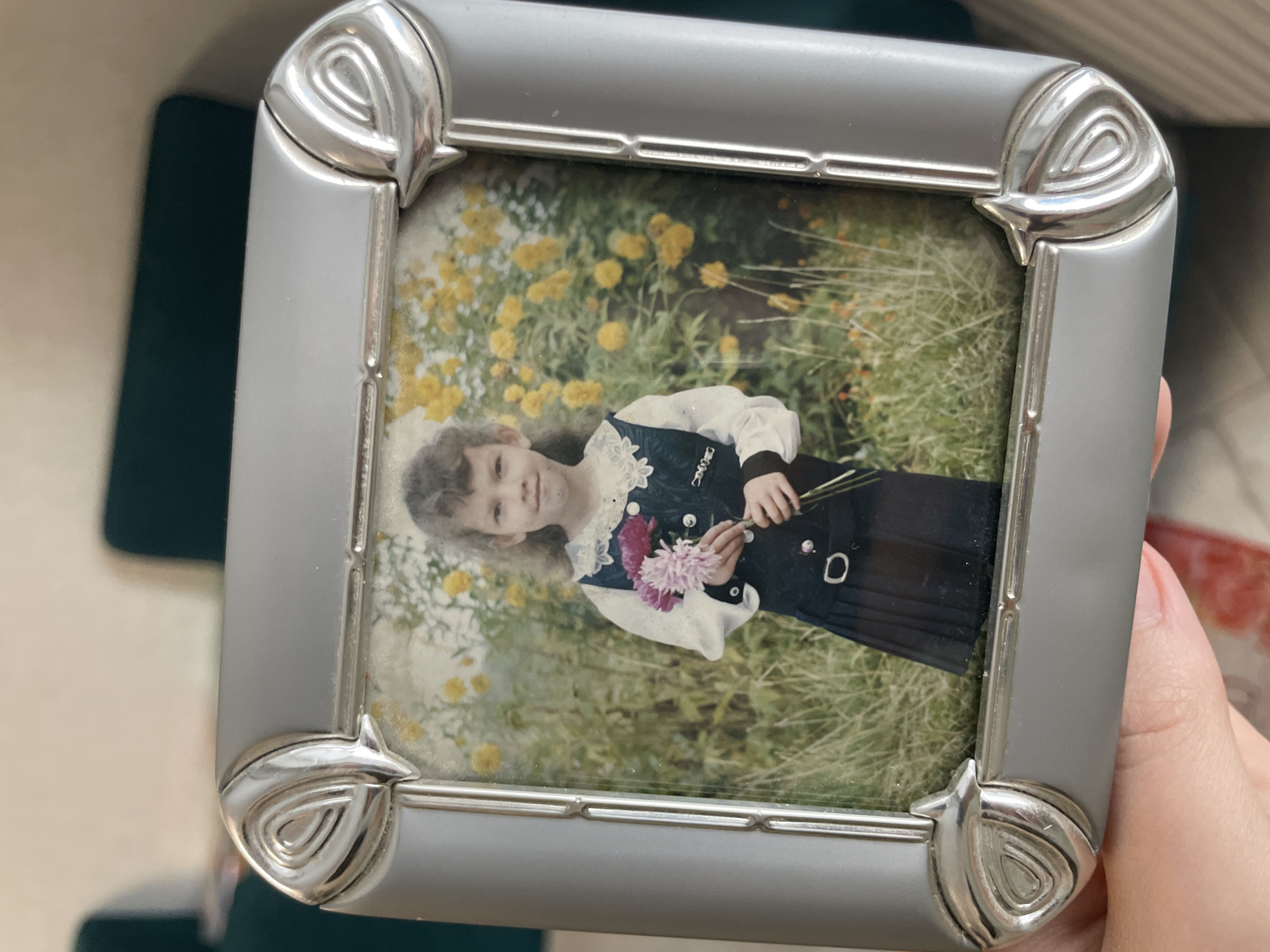 A photograph held by a hand features a framed picture of a young girl outdoors. The girl, wearing a dark dress with a white blouse and a lace collar, is holding a small bouquet of pink and purple flowers. She is standing amidst yellow wildflowers and tall green grass. The frame is silver, with a decorative design at each corner. There is no indication of the picture being taken in Izhevsk, Russia.