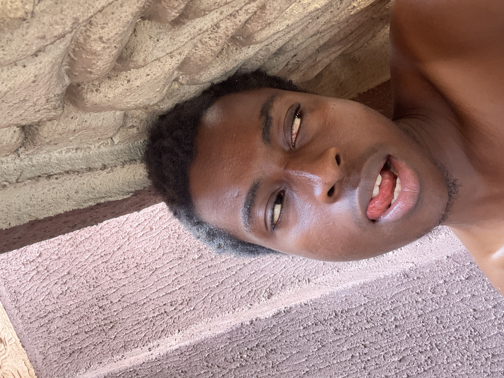 This close-up portrait, oriented diagonally, features a young person with dark skin and short, dark, curly hair. The individual's face is prominent, with one eye visible looking upward and to the side, and the mouth open with the tongue partially extended, conveying a playful or expressive look. Bright highlights are visible on the forehead, cheek, and bare shoulder, suggesting strong, possibly natural lighting.
The background consists of two distinct textured surfaces. The upper left portion shows a light brown or tan material, likely rough concrete or stone, characterized by thick, horizontal ridges. The lower right background is a lighter, pinkish-tan textured surface, possibly stucco or concrete, with fine, dense grooves or scratches. Shadows fall across these surfaces, indicating a strong light source. The setting appears to be an indoor or covered space with unfinished or roughly textured architectural elements. There is no visible text.