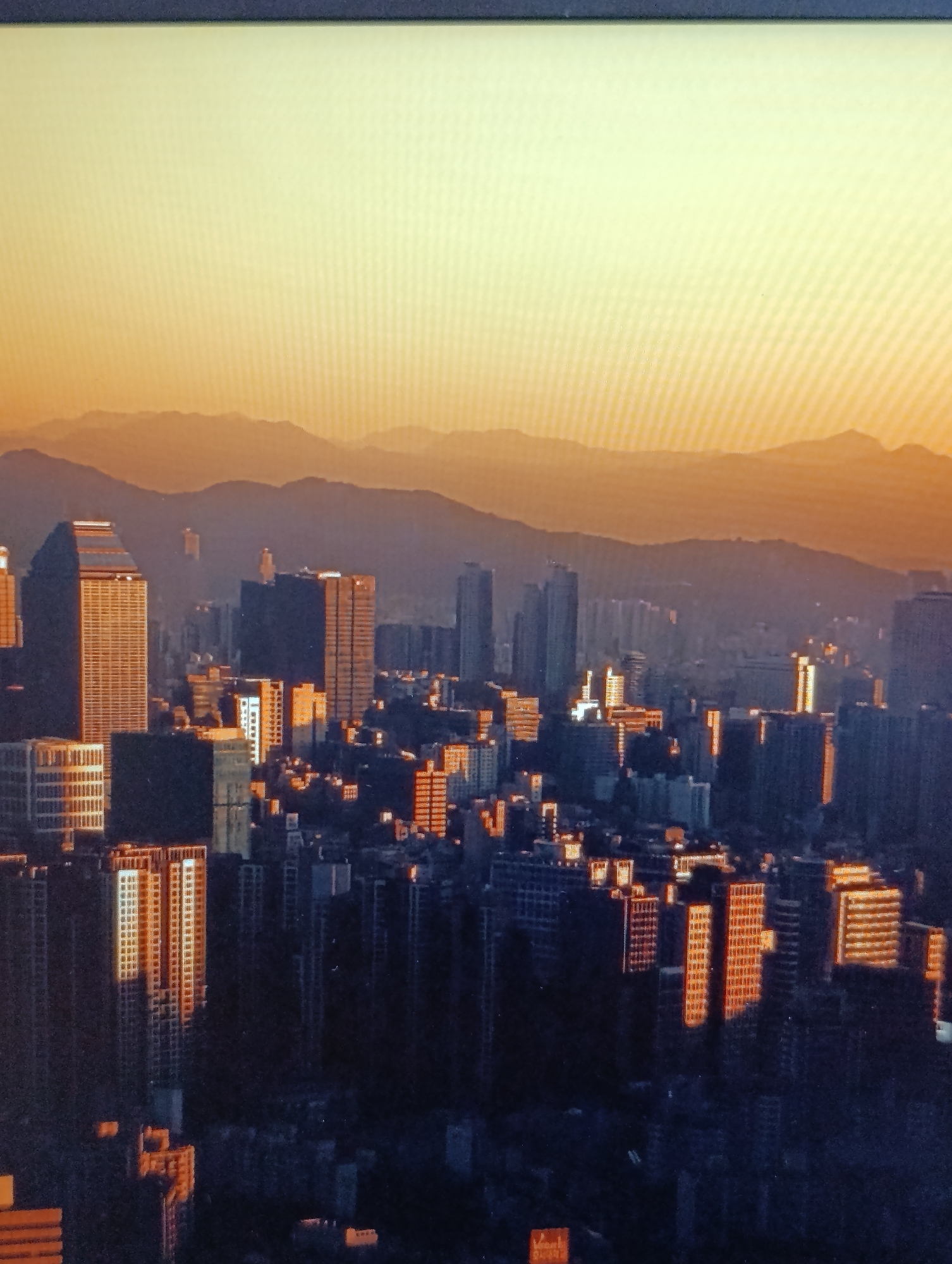 The image displays a panoramic view of a dense urban landscape at sunset. The sky is a gradient of warm yellow and orange, transitioning to a hazy horizon where distant, layered mountains are silhouetted. The city is characterized by numerous high-rise buildings, primarily composed of glass and concrete. The setting sun casts a warm, orange glow on the facades of some buildings, highlighting their architectural details. Shadows are long, and the overall atmosphere is one of twilight. The image does not contain any visible people or specific recognizable landmarks that would definitively place it in Dubăsari town council, Moldova. The focus is on the cityscape and the interplay of light and shadow.