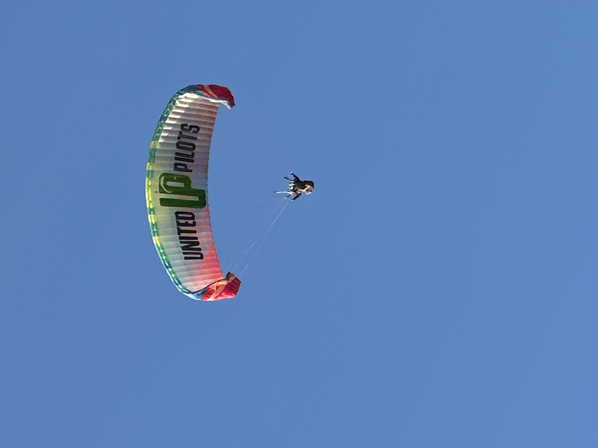 A color photograph captures a tandem paragliding event against a cloudless blue sky. The paraglider, curved significantly, displays the inscription "UNITED UP PILOTS" in bold green letters, with the words stacked vertically. The canopy's edges exhibit a striped pattern, transitioning from red to green. Two figures are suspended beneath the paraglider. The photograph was taken outdoors, during daylight hours. The setting is Fethiye, Turkey.