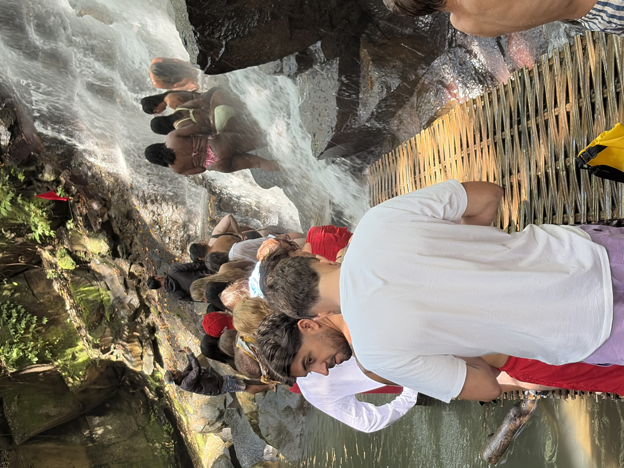 The image depicts a group of tourists gathered around a waterfall in Gianyar, Indonesia. The foreground shows several individuals standing on a woven bamboo walkway that spans across the water. One man in a white t-shirt and red shorts is prominently featured from the back, facing away from the camera. Another man, in a white long-sleeved shirt and red shorts, is visible to his left, looking towards the waterfall.
Behind them, a queue of people, mostly women in swimwear, are waiting to access the waterfall. Some are already in the water, being showered by the cascading falls. The waterfall itself is a multi-tiered cascade over moss-covered rocks, with lush green foliage visible on the surrounding cliffs. Sunlight filters through the trees, illuminating parts of the scene. A red flag is planted on the rocky incline to the left of the waterfall. The water appears clear and inviting, and the atmosphere suggests a popular tourist destination for swimming and recreation. The presence of a yellow bag on the bamboo walkway indicates personal belongings being carried by visitors.
