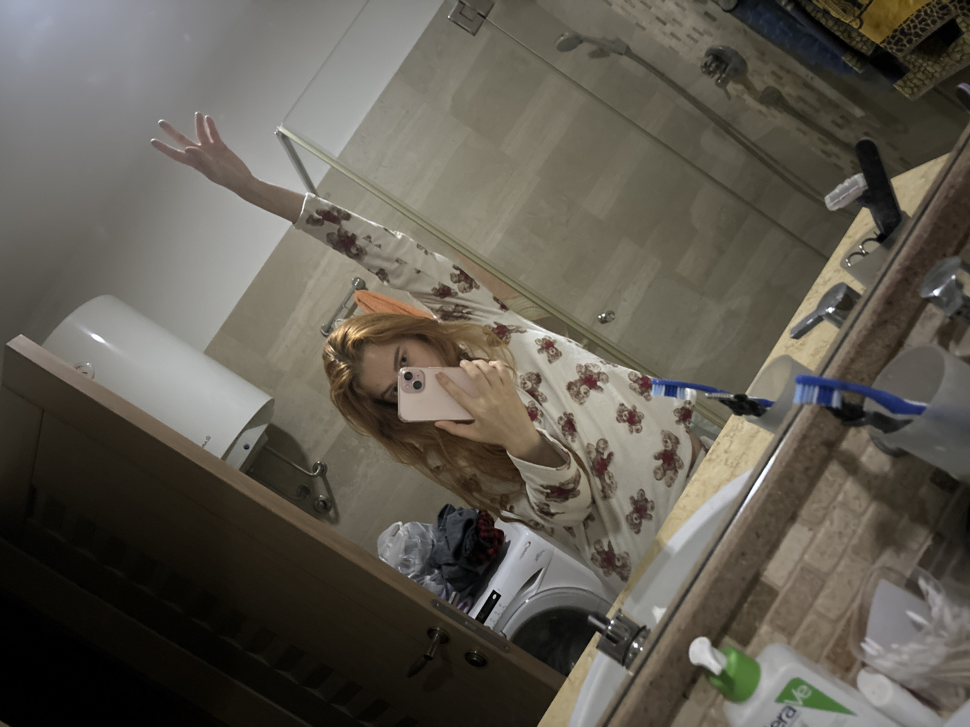 A fair-skinned, red-haired woman in bear-print pajamas is taking a selfie in a bathroom. She is holding a pink smartphone. Her left arm is raised above her head. The bathroom contains a shower with glass doors, a sink with various toiletries including two toothbrushes, and a white washing machine. A white cylindrical water heater is mounted on the wall. The location is likely in Budva, Montenegro, inferred from the architectural style and common bathroom fixtures.
