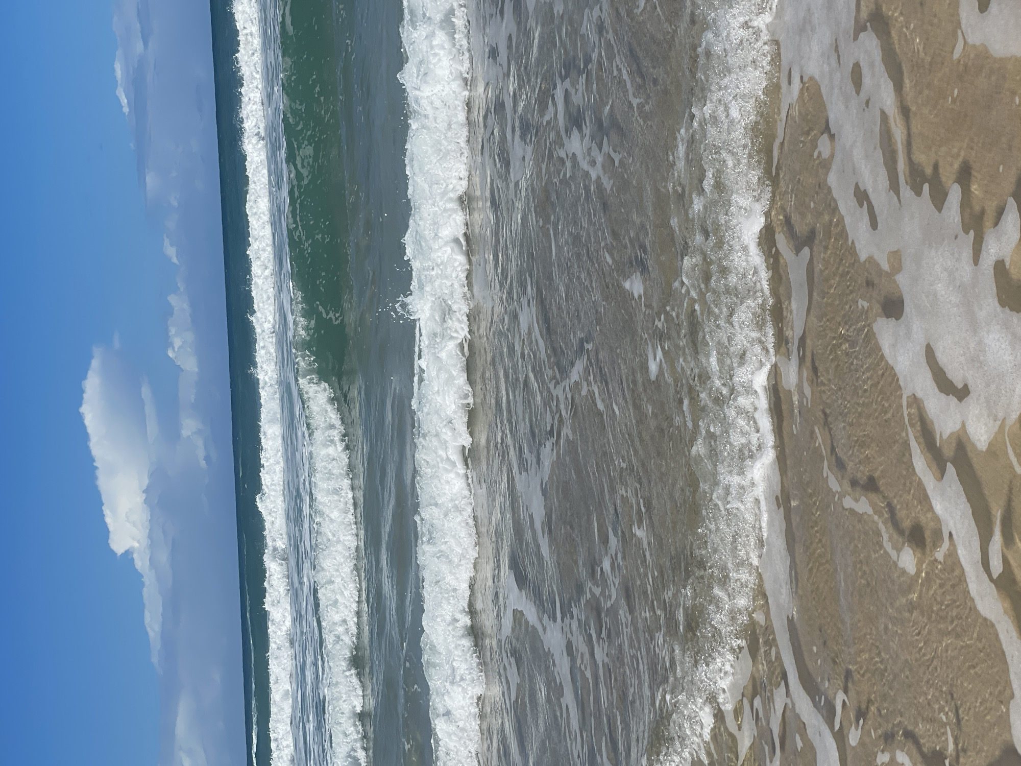 The image depicts a view of the ocean from a beach on the Gold Coast, Australia. Multiple waves are rolling towards the shore, with white foam cresting on their tops. The water is a clear turquoise green. The sky is blue with scattered white clouds. The sand in the foreground is wet and appears to be shallow water lapping at the shore. There are no people or man-made objects visible in the frame.