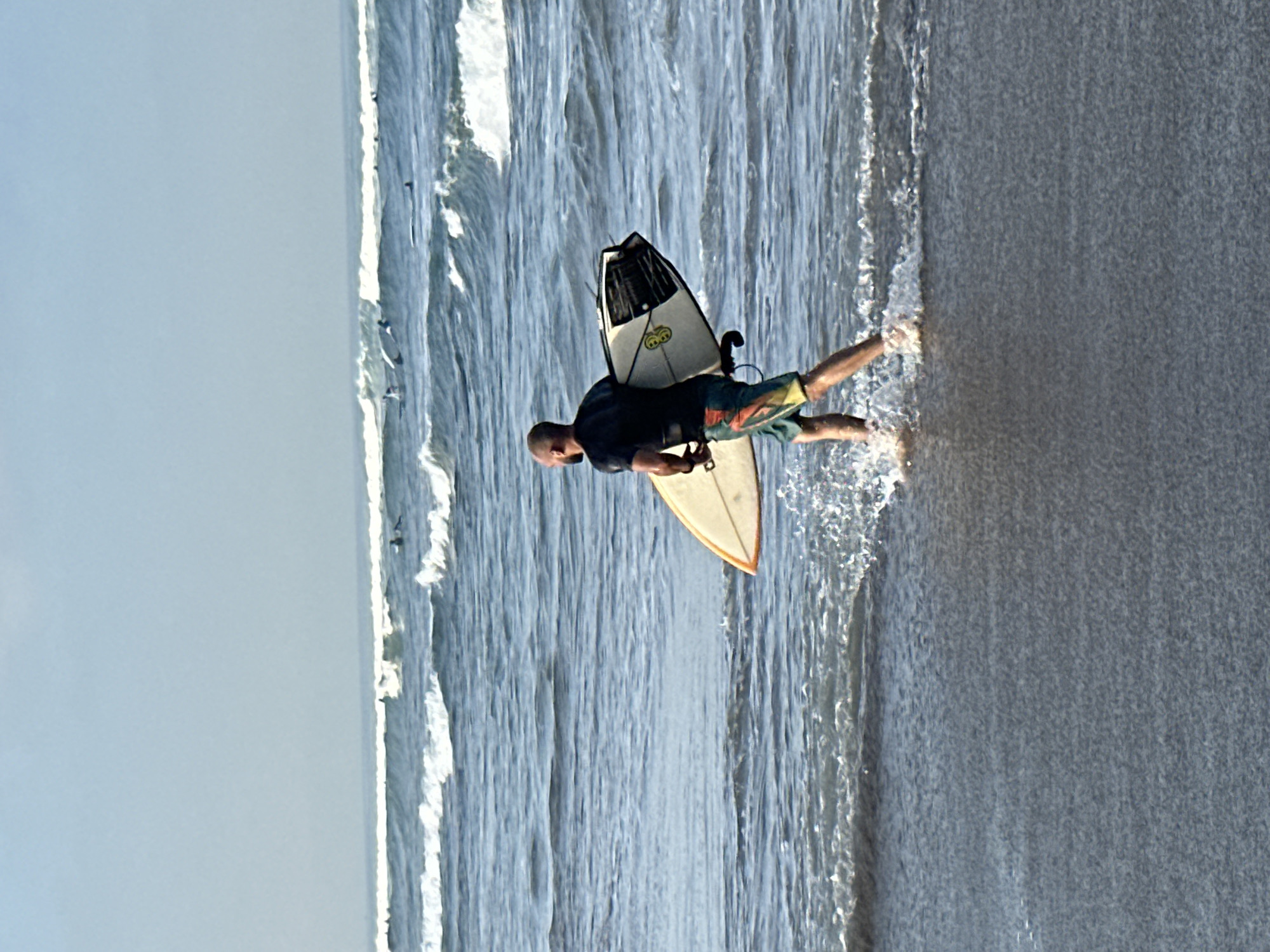 A bald man, wearing a black t-shirt and multi-colored swim trunks, walks through shallow ocean water while carrying a surfboard under his right arm. The surfboard is light-colored with an orange trim, and the back of the board features a black and white design with a yellow circular logo. The man is facing away from the camera, looking towards the ocean. He is in the process of entering the water, with small waves breaking around his legs and splashing around the surfboard. In the background, the ocean stretches out to the horizon with several other surfers visible in the distance. The sky is a clear, pale blue. The scene appears to be a beach in North Kuta, Indonesia, a popular surfing destination.