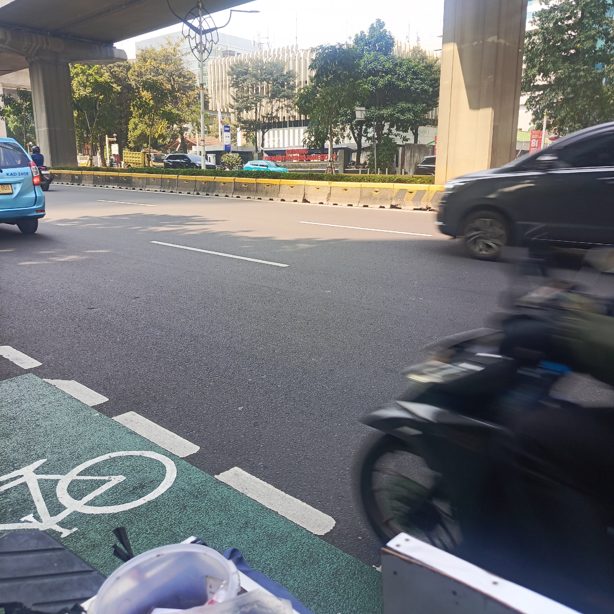 The image depicts a street scene in South Jakarta, Indonesia, likely in the Kuningan area due to the architecture visible in the background. An elevated road structure, supported by concrete pillars, looms overhead, casting shadows on the asphalt road below. The road features a dedicated bicycle lane painted in green with a white bicycle symbol.
In the foreground, a portion of a motorcycle, blurred due to motion, is visible, indicating movement. A blue car with license plate "KAD 2408" is parked on the left side of the road. The background shows a modern building with vertical louvers, suggesting an urban business district. Trees line the street between the road and the building. Traffic barriers, made of concrete segments with yellow markings, divide the road. The scene suggests a typical day in a bustling urban environment.