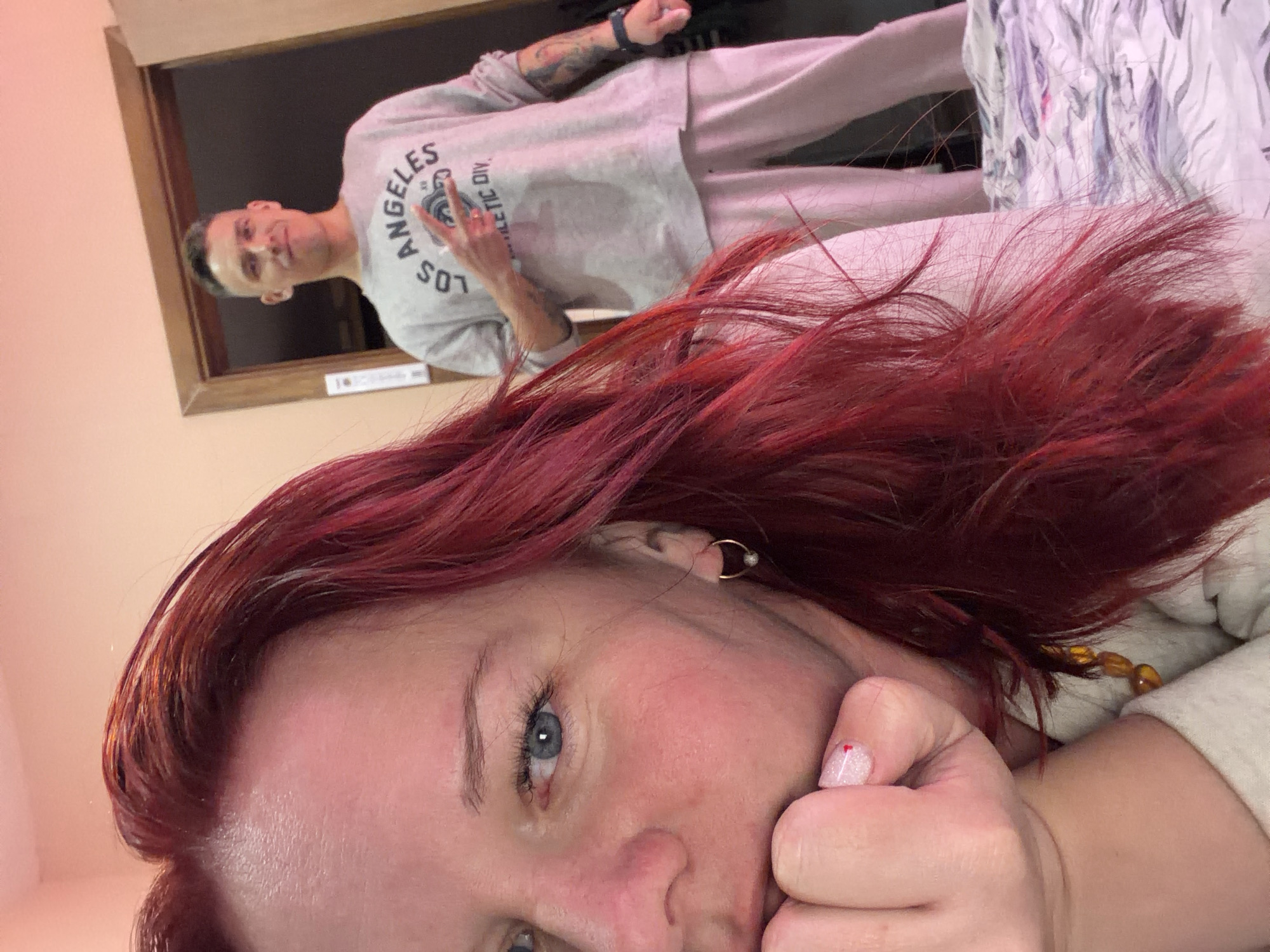 This image depicts an indoor residential scene in Chaikovsky, Russia. In the immediate foreground, a Caucasian female with bright red hair and blue eyes is visible, resting her chin on her right hand. Her fingernails are painted white with a small red heart accent on the thumb. She wears a small gold-colored hoop earring with a grey bead in her visible ear and amber-colored beads around her neck. Her gaze is directed towards the camera. She is situated on a bed covered with white, purple, and grey patterned bedding. In the background, framed by a dark brown wooden doorway, a Caucasian male stands, slightly smiling, and making a peace sign gesture with his right hand. He has a short, faded haircut, visible tattoos on his left forearm, and wears a dark watch on his left wrist. He is dressed in a light grey long-sleeved sweatshirt bearing "LOS ANGELES ATHLETIC DIV" text and light grey sweatpants. The walls of the room are painted a pale, warm tone.