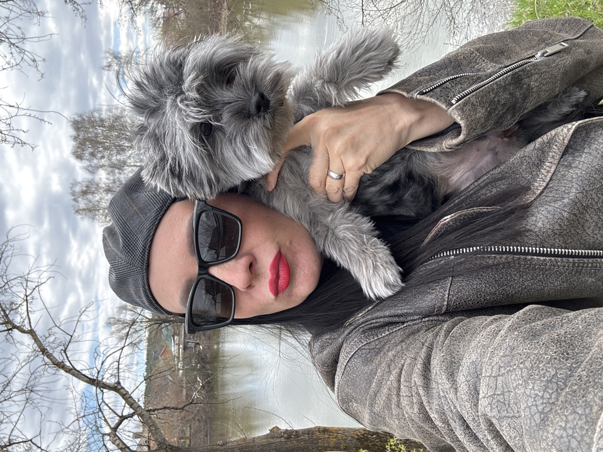 A person wearing a gray plaid cap and black sunglasses, with red lipstick, is holding a gray Schnauzer-type dog. The person is wearing a gray-brown leather jacket. The background includes a body of water (likely a river or lake), bare trees, and a cloudy sky. The location is most likely in Chaikovsky, Russia.