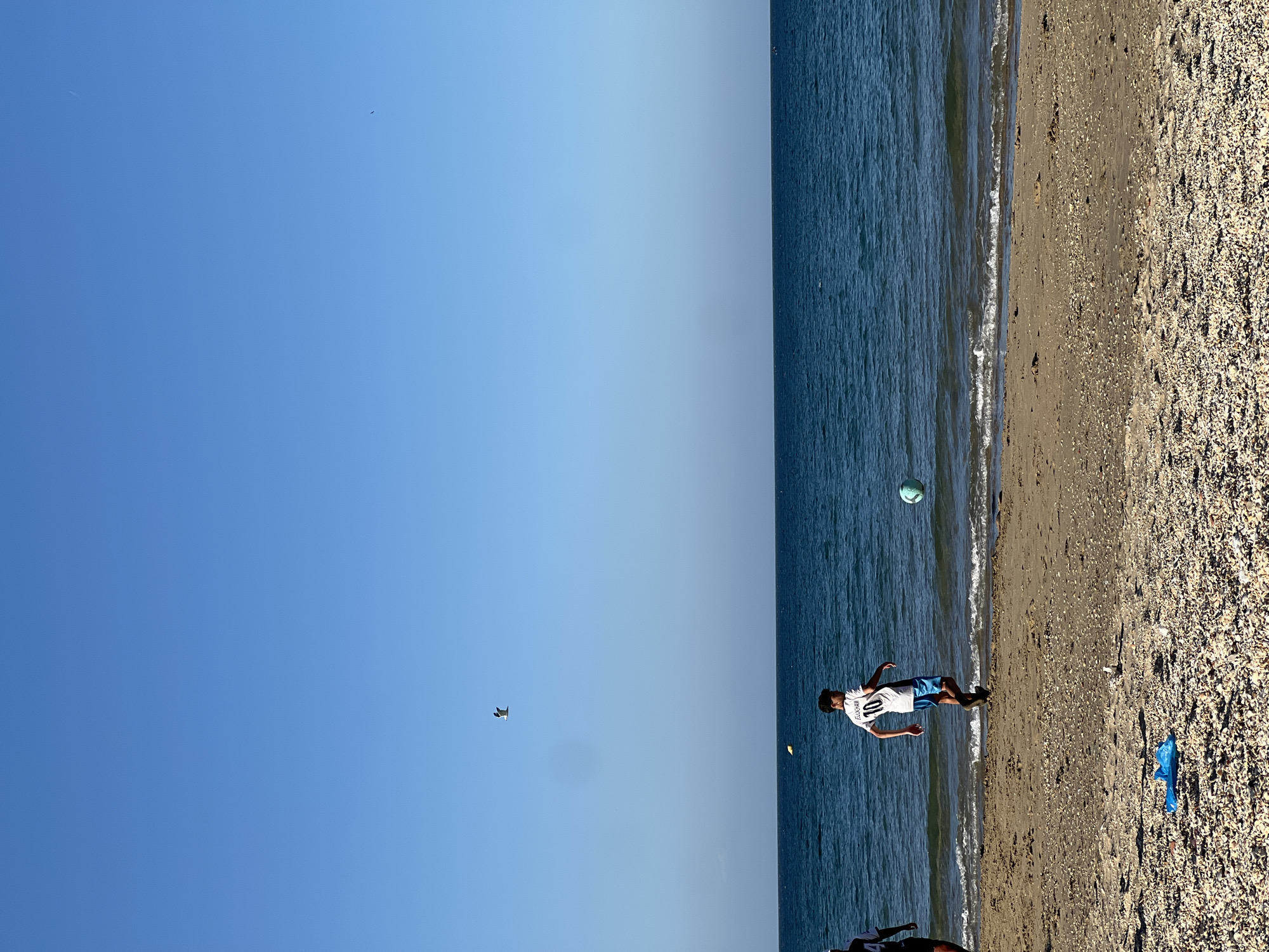 The image depicts a beach scene in M'Diq, Morocco. The sky is a clear blue. A boy wearing a white jersey with the number 10 and blue shorts is playing on the beach. A green soccer ball is airborne near the water's edge. A drone-like object is visible in the sky. The beach is sandy with shells. A blue plastic bag lies on the sand. Another person is visible in the bottom left.