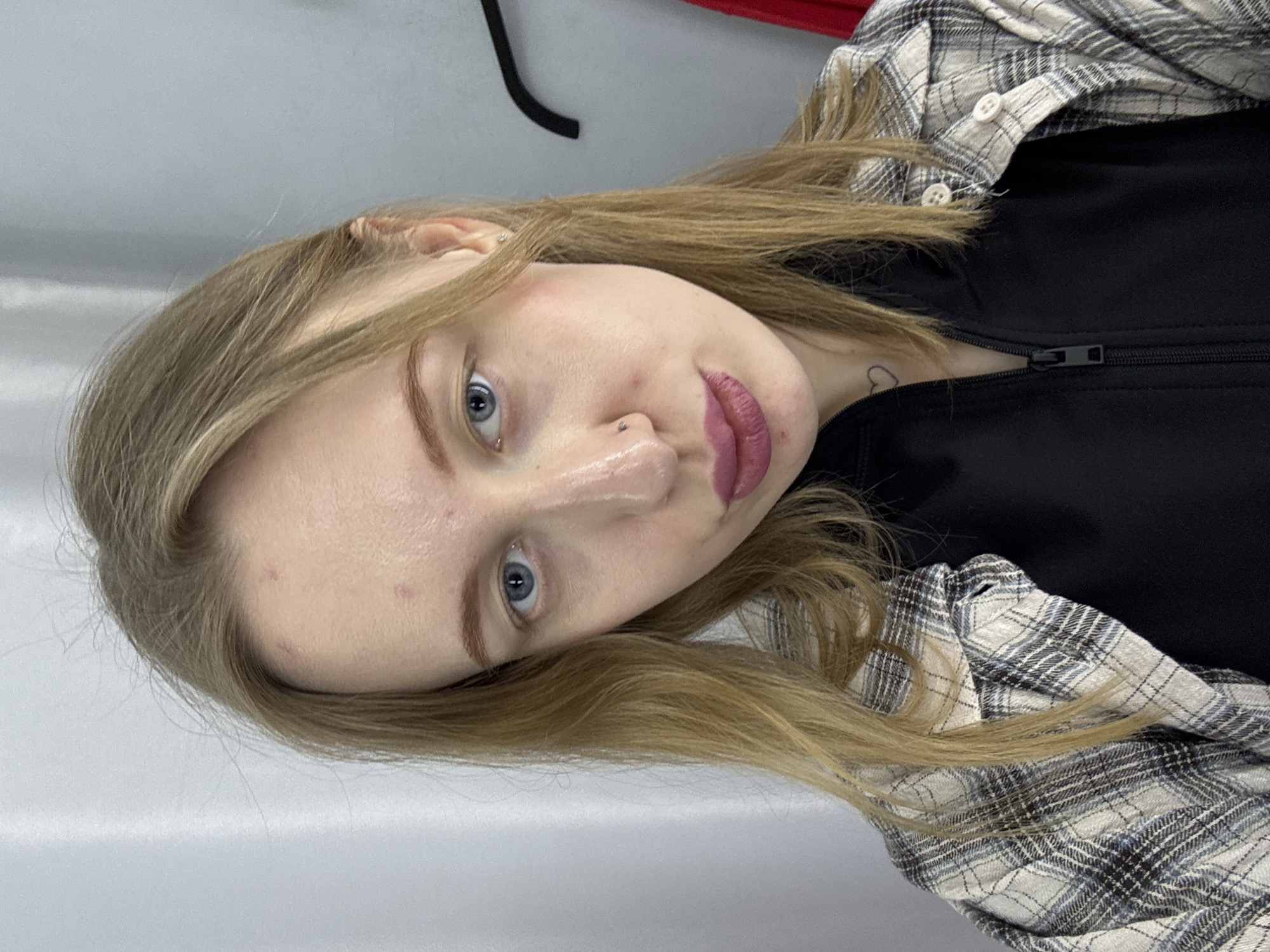 A portrait of a young woman with blonde hair and blue eyes. She is wearing a black zip-up jacket and a plaid shirt. She has a piercing on her nose and a small heart tattoo on her neck. The background appears to be a plain wall. There is no information available to determine the location in Rostov-on-Don, Russia.