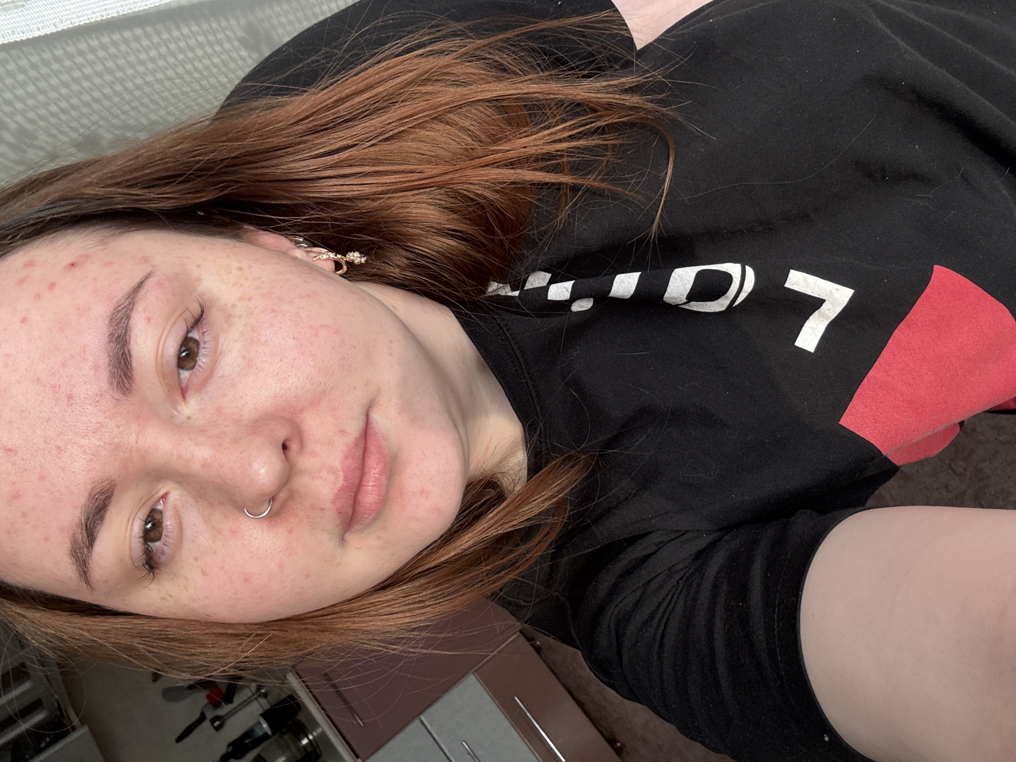A selfie taken indoors shows a young woman with auburn hair and light skin. She has a nose ring, freckles, and what appear to be acne on her face. She is wearing a black long-sleeved shirt with white and red graphic elements. Part of a room with cabinets and a window blind is visible in the background. There is no indication that the photo was taken in Izhevsk, Russia.