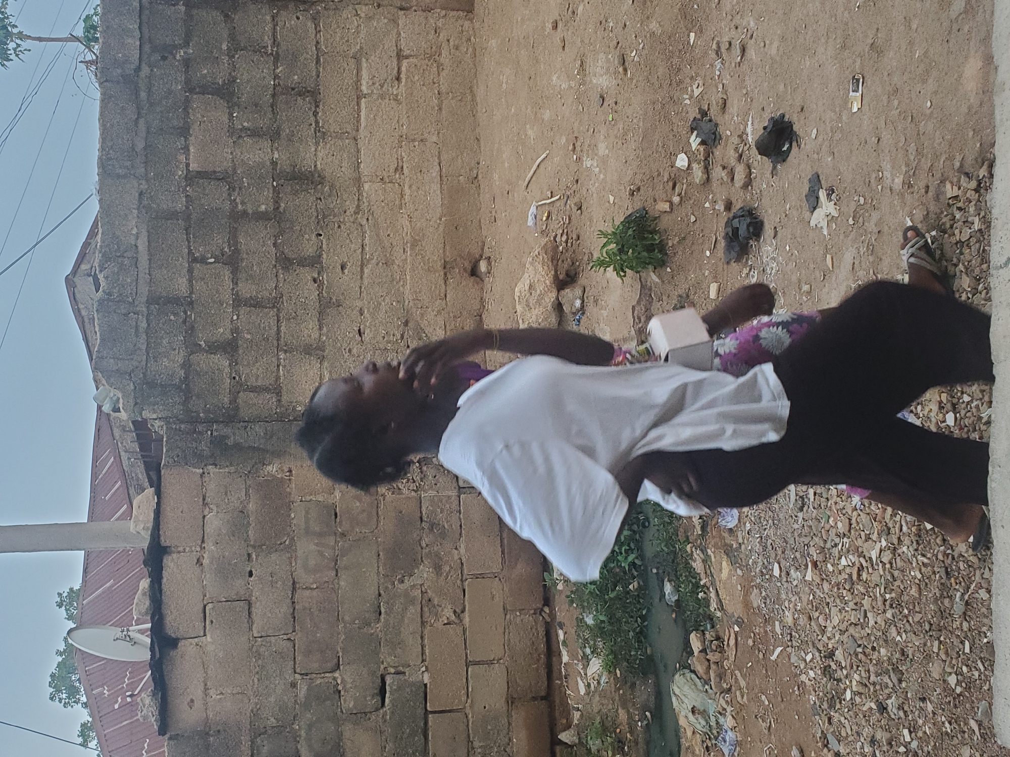 A person stands outside a brick building on a dirt and gravel lot strewn with trash. They wear a white t-shirt, dark pants, and sandals. A satellite dish is affixed to the building. A small drainage ditch with vegetation runs along the edge of the lot. The location is consistent with an urban environment in Jos, Nigeria.