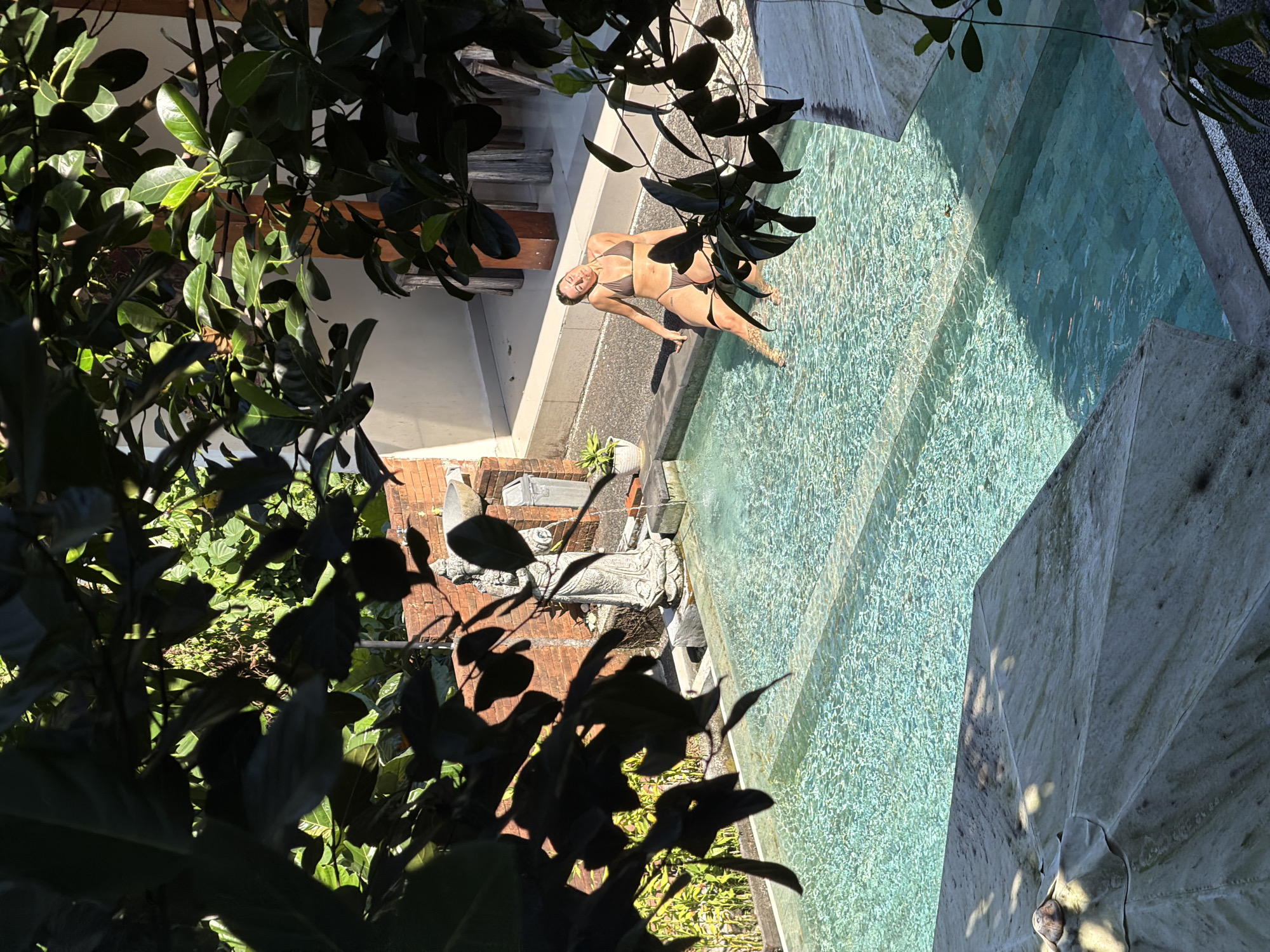 In Ubud District, Indonesia, a woman in a brown bikini sits on the edge of a turquoise swimming pool, her legs submerged in the water. She is leaning back with her head tilted upwards, eyes closed, basking in the sunlight filtering through lush green foliage. The pool is surrounded by a paved area and a brick wall, with a stone statue of a figure standing near the water's edge. The scene is framed by dark, leafy branches that cast shadows on the water and surrounding area, suggesting an outdoor setting, likely a private villa or resort.