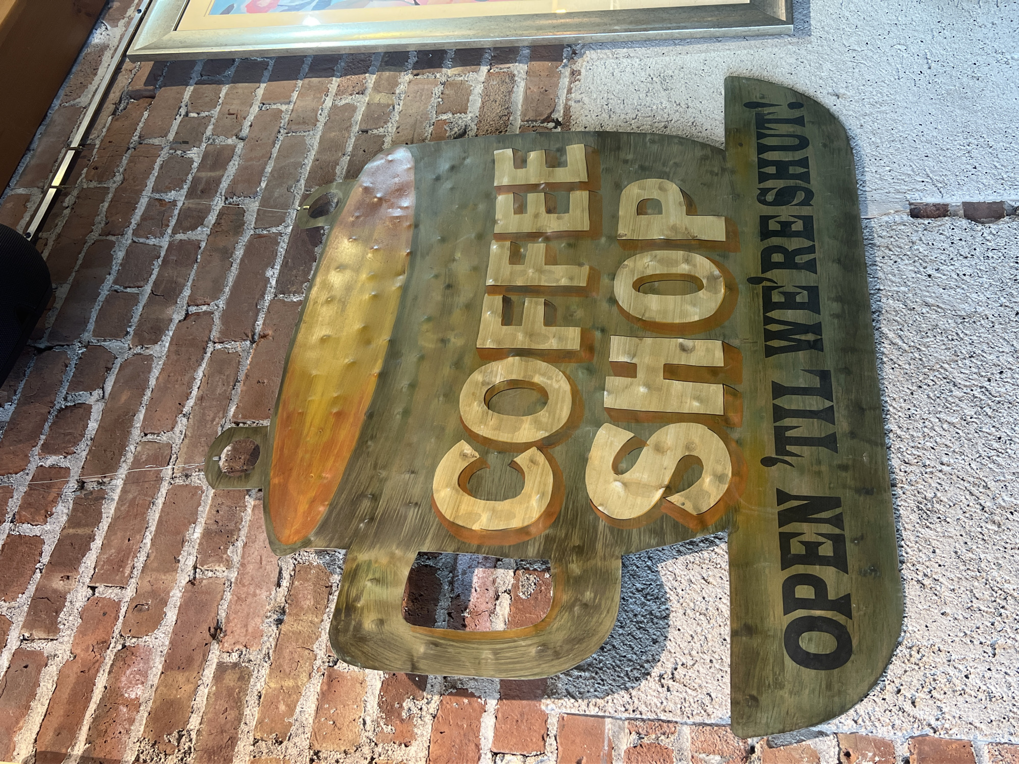 A vertically oriented, medium shot depicts a wooden sign with the words "COFFEE SHOP OPEN 'TIL WE'RE SHUT!" mounted on a brick wall and a textured wall.
The sign is shaped like a coffee mug. The word "COFFEE" is written vertically in large, gold, three-dimensional block letters. Below "COFFEE" is "SHOP," also in gold block letters. To the right of the mug shape, a wooden panel reads "OPEN 'TIL WE'RE SHUT!" in black lettering.
The brick wall behind the sign is red and aged, with white mortar between the bricks. The adjacent wall is textured and painted a light color. A framed picture hangs above the sign. The scene suggests an establishment in the City of Newburgh, United States of America.