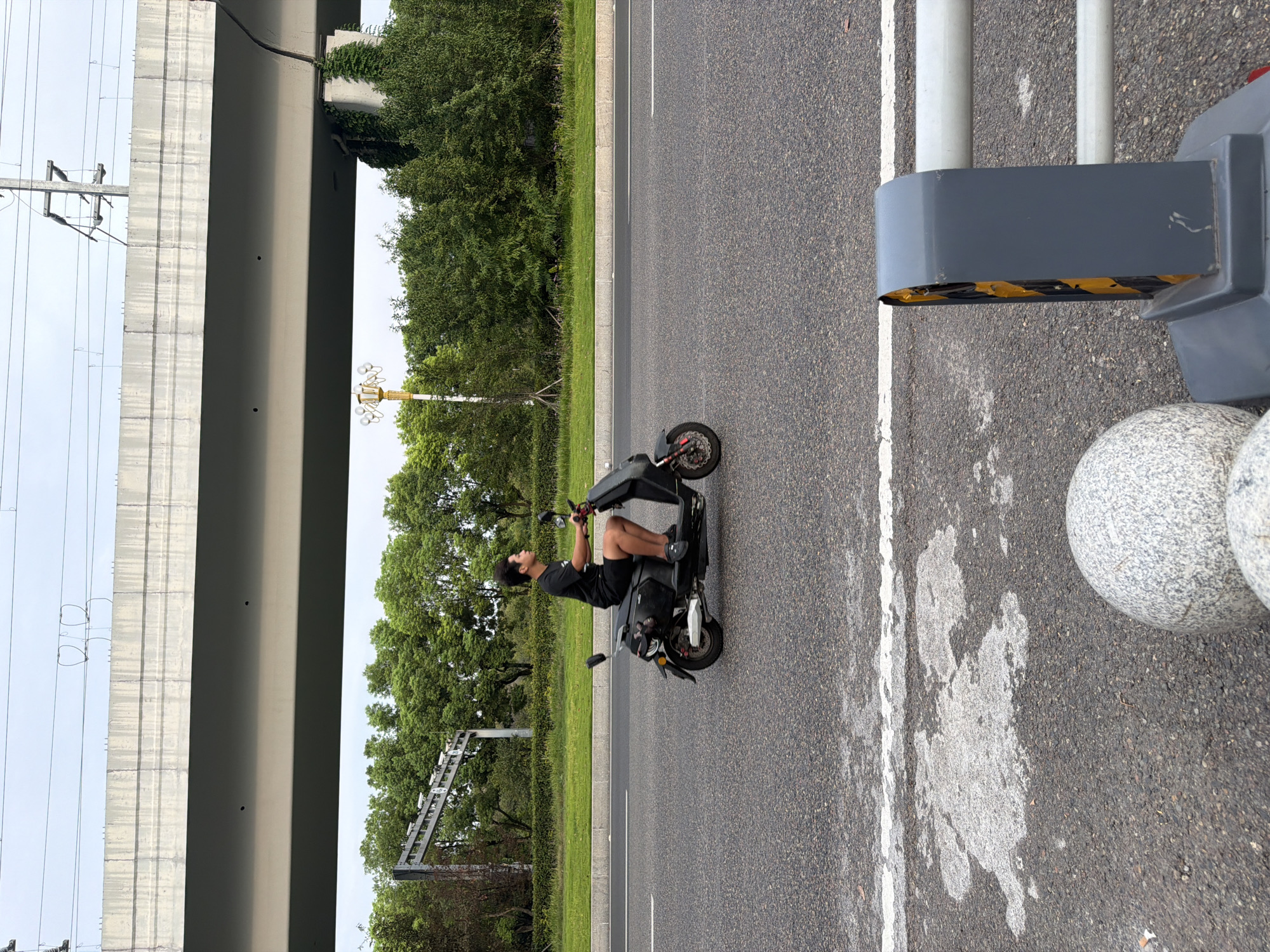A young man with dark, curly hair is riding a black electric scooter down an asphalt road in Jiangjin, China. He is wearing a black t-shirt and black shorts. The scooter has a dark gray body with some visible components. The road has a white line marking the lane. In the background, there is a large overpass, a line of green trees, and a utility pole with wires. The sky is overcast. In the foreground, there are two large, round granite bollards and a gray traffic barrier with yellow and black hazard tape.