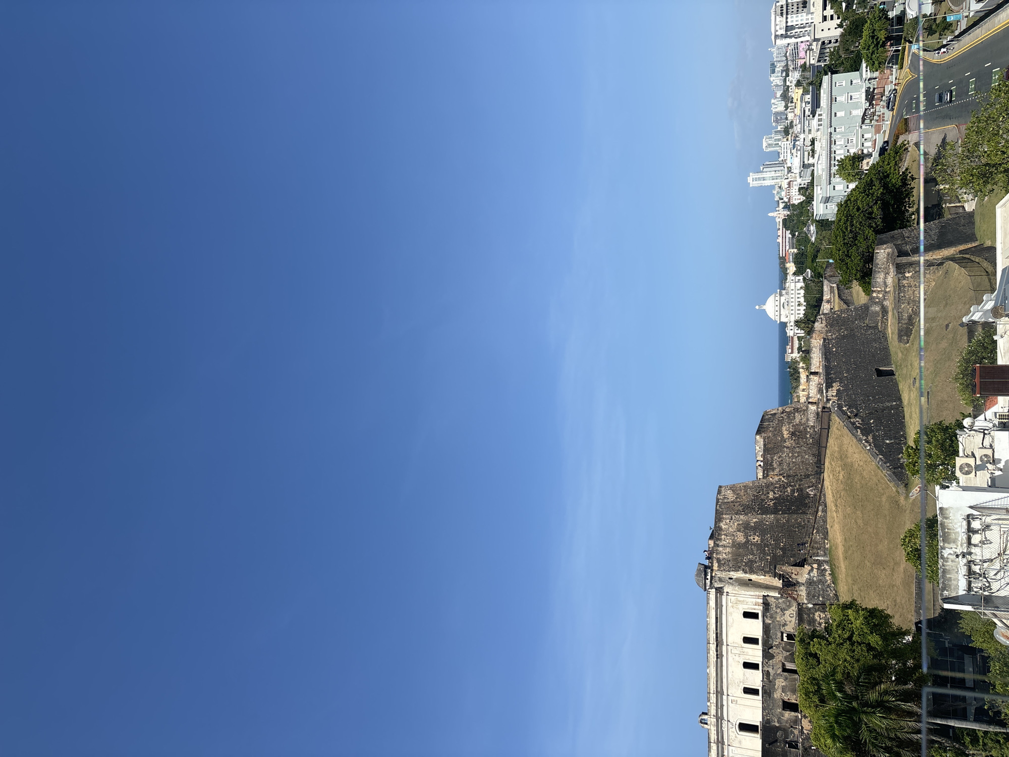 The image displays a panoramic view of San Juan, Puerto Rico, a U.S. territory. In the foreground, the imposing stone walls of Castillo San Felipe del Morro, a 16th-century Spanish fortress, dominate the left side of the frame. The fortress is characterized by its weathered, dark stone, battlements, and expansive green lawn leading up to its structure. Adjacent to the fortress, a cluster of palm trees and other lush vegetation adds a touch of tropical greenery.
Beyond the fortress, the cityscape of San Juan unfolds. A prominent white dome, likely belonging to a significant building like the San Juan Cathedral or the Capitol Building, stands out against the backdrop of modern high-rise buildings. The urban landscape features a mix of architectural styles, with some older colonial-era structures and more contemporary buildings. A paved road with cars is visible in the mid-ground, indicating the presence of active city life. The scene is bathed in bright daylight under a clear, expansive blue sky, suggesting fair weather conditions. The overall impression is one of a historic city meeting modern development, with the ancient fortress serving as a stark reminder of its past.