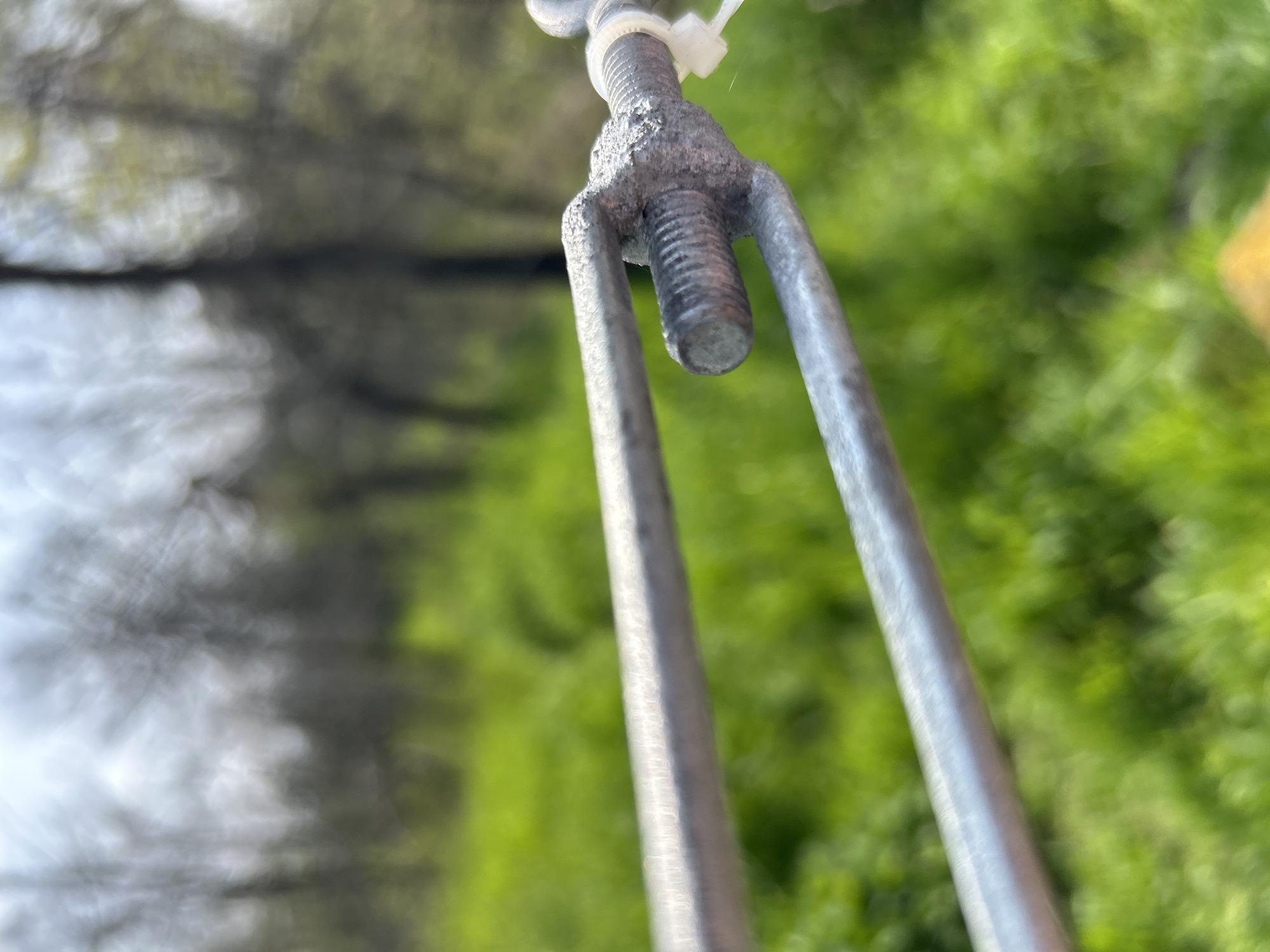 Close-up, upward shot of a metal turnbuckle, secured with a white cable tie, suspended in front of a blurred background of greenery and out-of-focus trees. The location cannot be determined.
