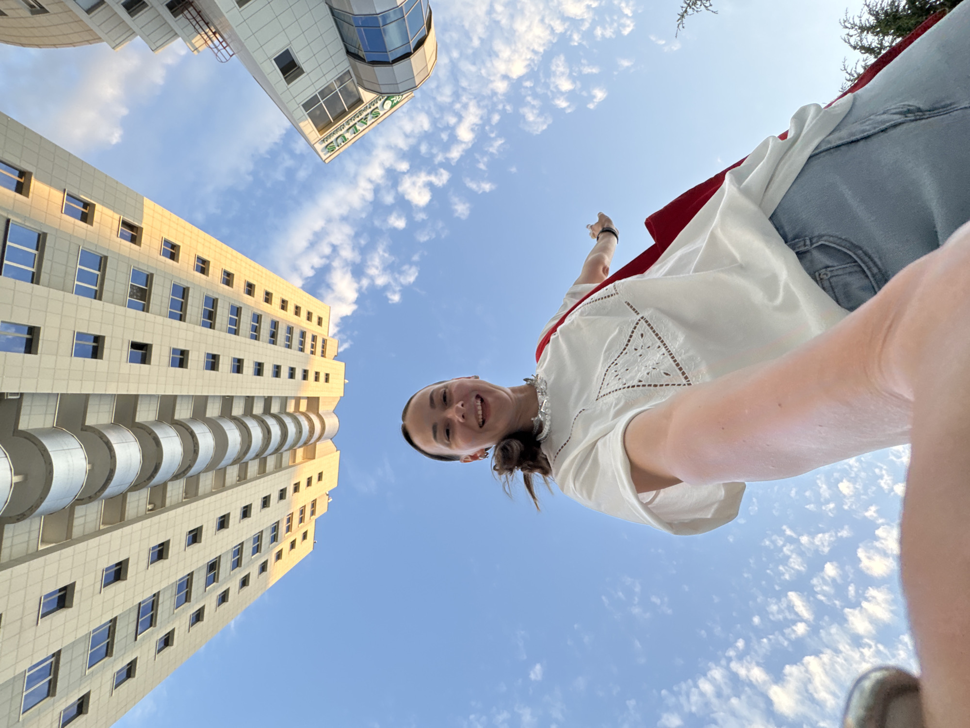 An upward-angle, low-resolution, full-shot photograph taken from a low perspective shows a young woman standing in front of two buildings under a clear blue sky filled with scattered clouds. The woman, positioned in the center of the frame, is smiling and looking at the camera. She has her left arm extended towards the camera, and her right arm is bent at the elbow, with her hand partially visible. She is wearing a white shirt with lace detailing on the neckline and sleeves, light blue jeans, and a red scarf draped over her shoulders. The buildings appear to be modern high-rises. The building on the left is tall and rectangular with a beige facade and balconies. The building on the right is shorter, with a white facade and a curved section. A sign on the right building reads "CASALUX". The lighting suggests it is daytime, with sunlight illuminating the buildings. The context provided indicates the location is Sochi, Russia, specifically near a building with the "CASALUX" signage.