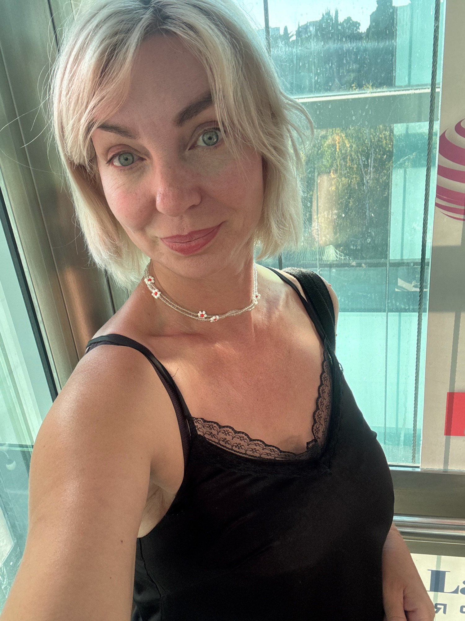 A fair-skinned woman with blonde hair is taking a selfie in front of a window in Sochi, Russia. She is wearing a black camisole with lace trim and a beaded necklace with small red and white flowers. The window shows a blurred view of trees and buildings. A portion of a sign with pink and white stripes is visible on the right side of the frame.