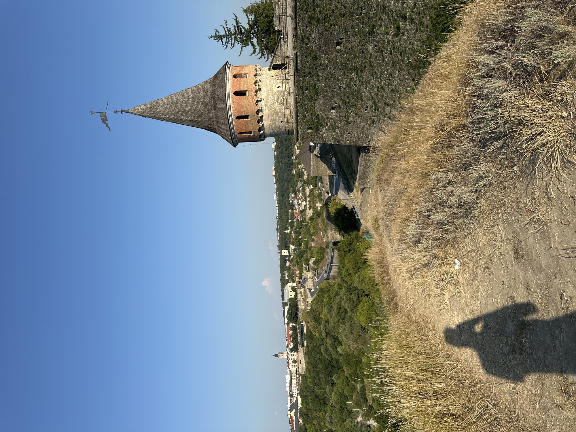 The image displays a view of the Kamianets-Podilskyi Castle in Kamianets-Podilskyi, Ukraine, on a clear day. In the foreground, a dirt path is visible with dry grass and vegetation on either side. A shadow of a person holding a phone is cast on the path, indicating a selfie is being taken. To the right, a large stone wall of the fortress is prominent, topped by a circular brick tower with a conical wooden roof, a flag, and a weather vane at its peak. Beyond the castle walls, a cityscape unfolds with numerous buildings, including churches with spires, nestled amongst green trees and rolling hills. A road with some vehicles is visible winding through the landscape. The sky is a vivid, cloudless blue.
