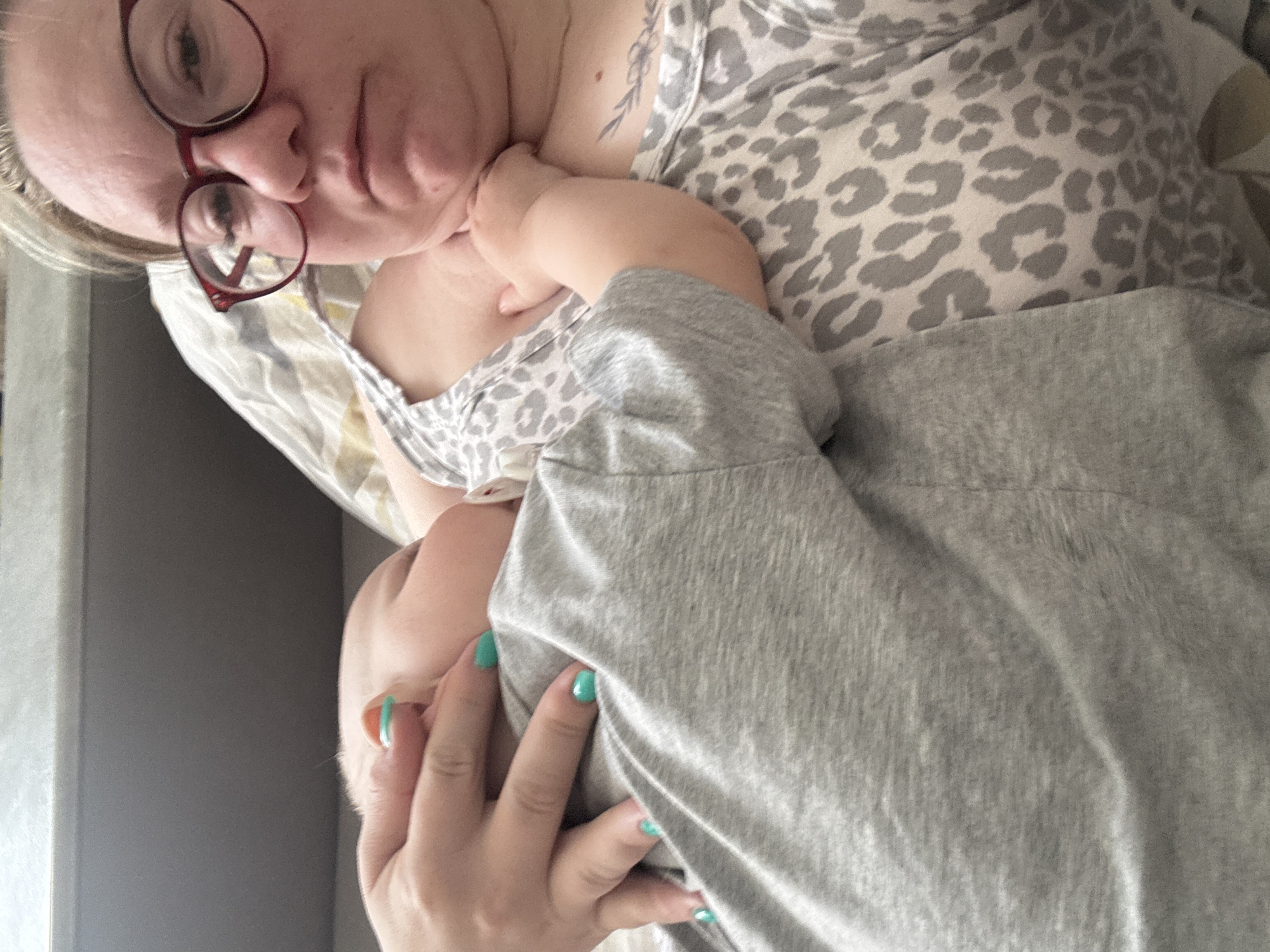 A woman wearing eyeglasses is lying down in a bed, dressed in a gray and white patterned tank top. Her right arm is draped over a small child dressed in a gray shirt. The woman’s left hand, with painted turquoise fingernails, rests on top of the child. The background is blurry, but a window with gray surroundings is visible. The location cannot be determined as Chaikovsky, Russia.