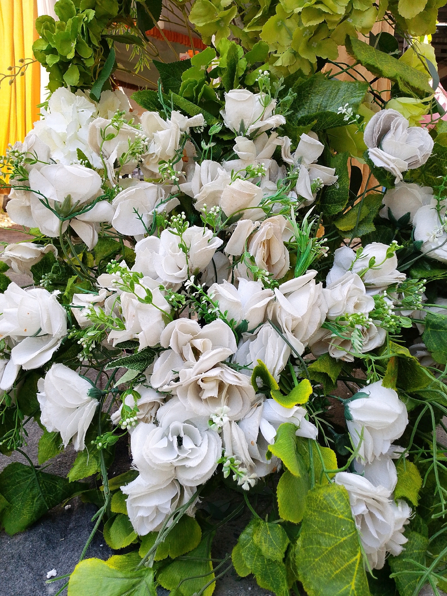 A close-up, eye-level shot captures an arrangement of artificial white roses interspersed with artificial green foliage. The roses appear to be made of fabric, with some showing hints of off-white or beige tones. Small, delicate white buds or accents are scattered among the roses and leaves. The background features hints of bright yellow fabric on the left and more muted, light brown fabric or material behind the floral arrangement. The ground beneath the arrangement is a dark, textured surface, possibly stone or concrete. The lighting is even, suggesting an indoor or shaded outdoor setting. There are no people or discernible actions in the image. The location is the city of Sugio, Indonesia.