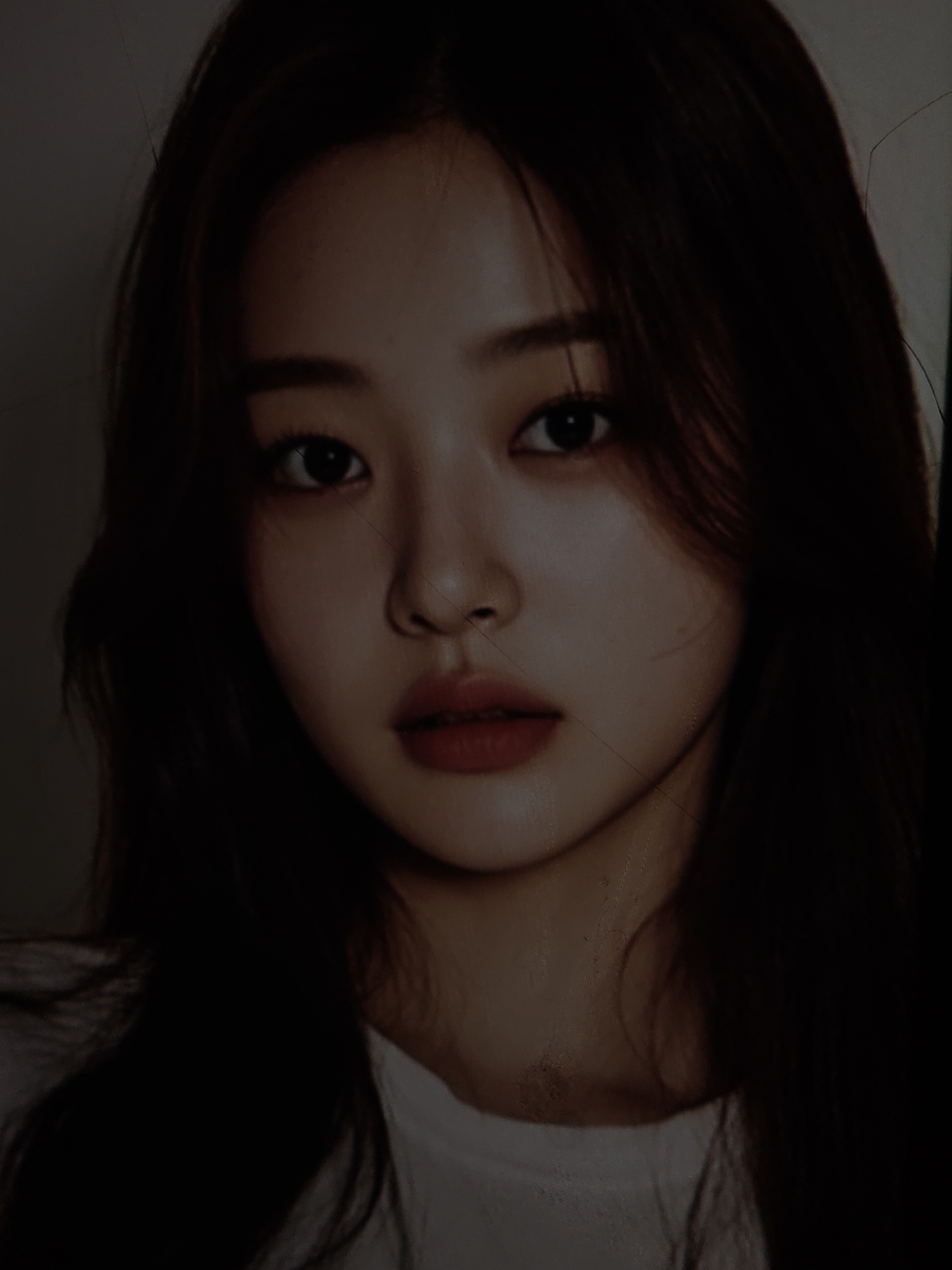The image is a close-up portrait of a young woman with long, dark hair. She is looking directly at the camera with a neutral expression. Her makeup includes defined eyebrows, eyeliner, and red lipstick. The lighting is somewhat dim, with shadows obscuring parts of her face and hair. There appear to be subtle linear imperfections on the surface of the image, possibly scratches or dust. The background is plain and out of focus. The scene does not contain any elements that would indicate a specific location within the city of Kurganinsk, Russia.