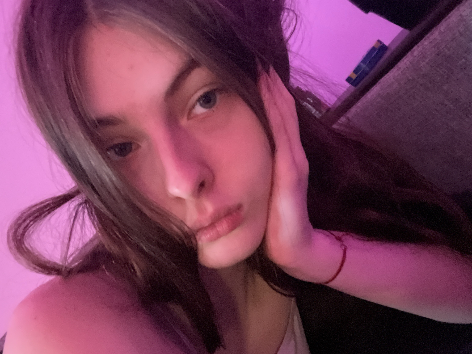 A young woman with long brown hair and light eyes is seen indoors, likely at night, given the purplish ambient lighting. She is resting her face on her hand, with a thin red bracelet visible on her wrist. A couch and a small blue object are partially visible in the background. The specific location is not discernible from the image, and no information suggests it is in Saint Petersburg, Russia.