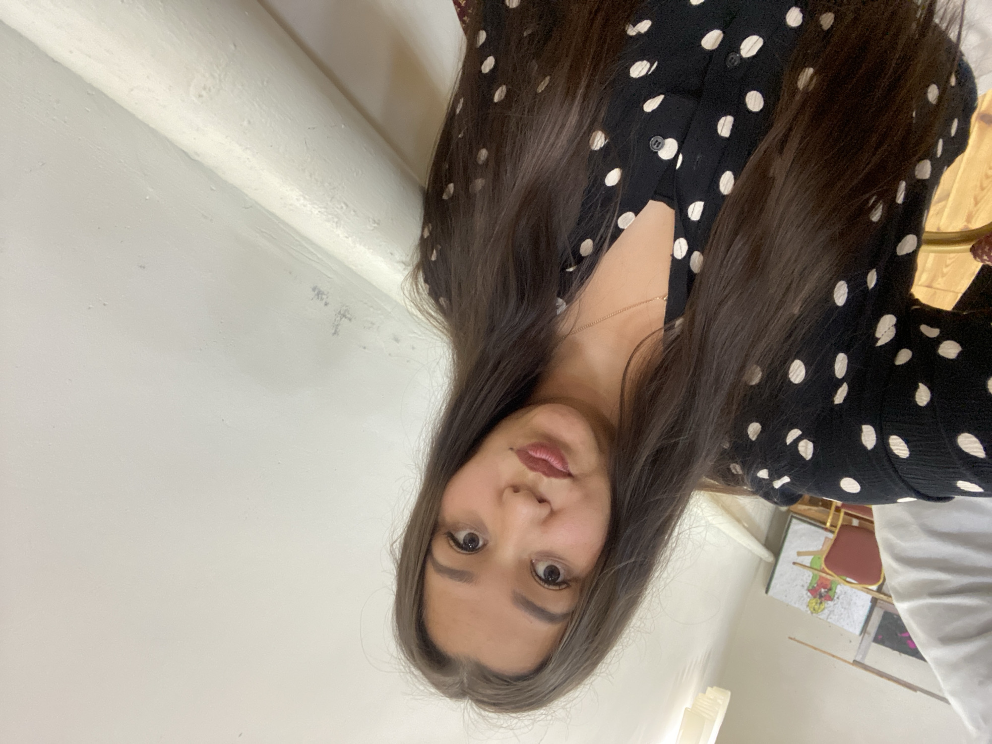 In Maloarslangulovo, Russia, a woman with long, dark brown hair and a polka-dot blouse poses for a selfie indoors. She is positioned centrally in the frame, with her face slightly angled towards the camera. The background consists of a plain, light-colored wall and a white pipe running along it. To the left, a portion of a white object, possibly a piece of furniture or clothing, is visible, along with a glimpse of an art piece on the wall depicting a figure. The lighting appears to be artificial, illuminating the scene evenly.