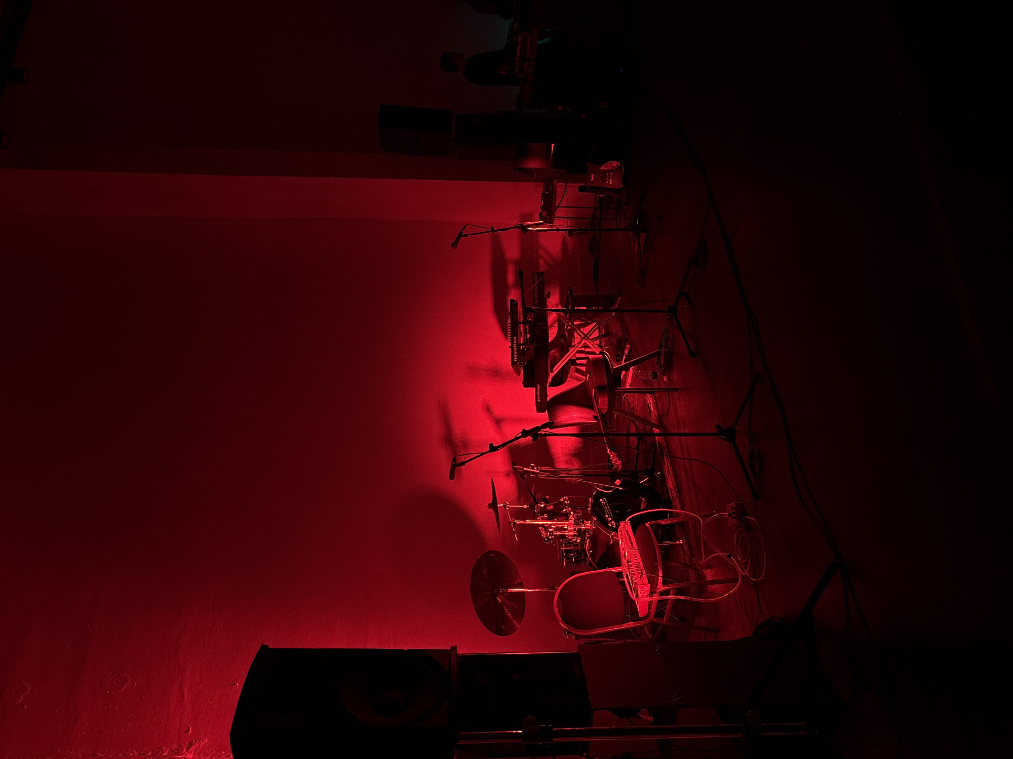 The image depicts a dimly lit room, possibly a performance space or studio, bathed in red light. On the wall, there is an assortment of musical instruments and equipment, including drum kits, microphone stands, and cables, all seemingly mounted or suspended. In the foreground, a large speaker cabinet sits on the floor. The setting is likely a stage or rehearsal area. Location cannot be determined from the image.