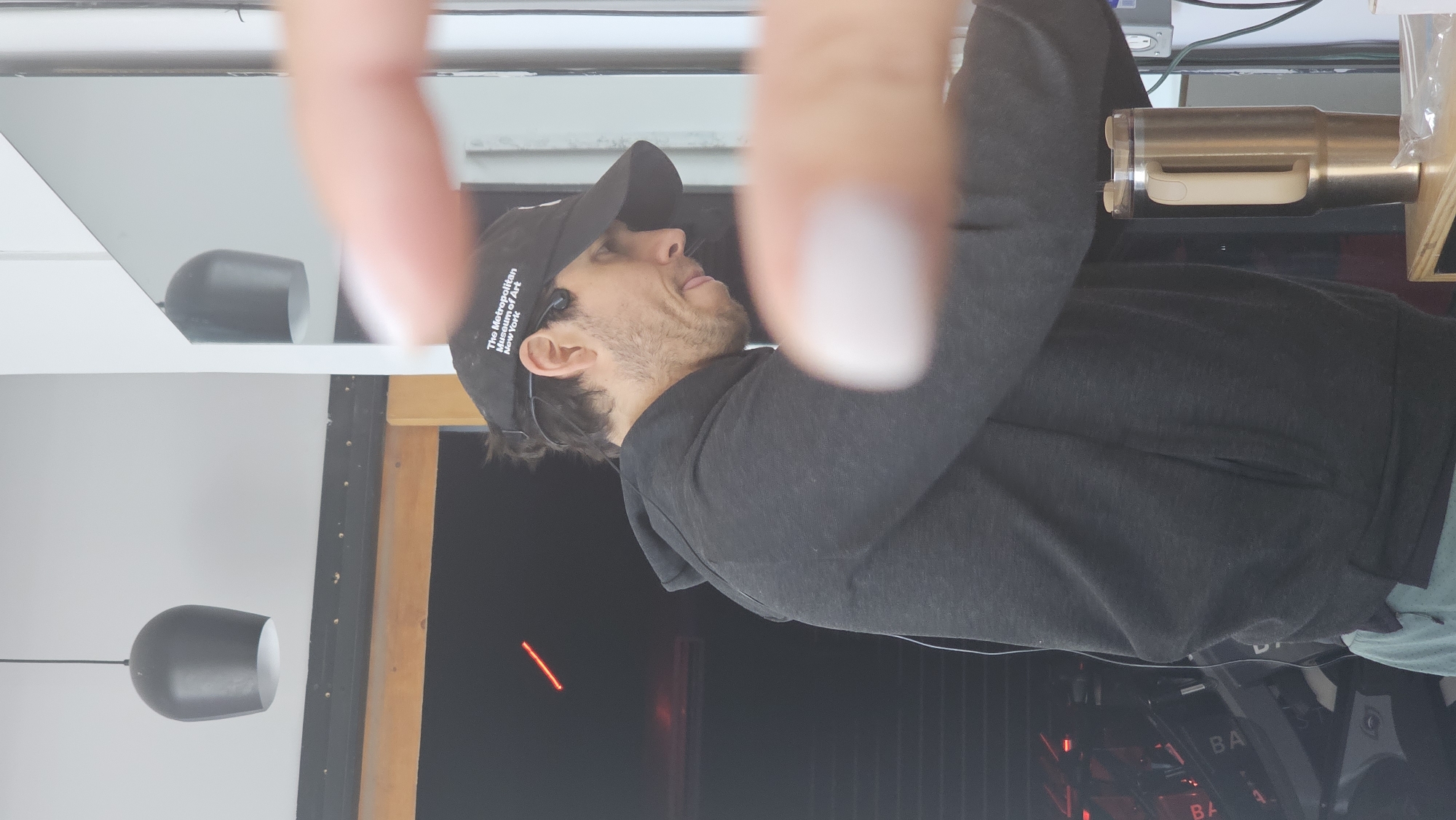 A man with a beard is wearing a black hoodie and a black baseball cap that reads "The Metropolitan Museum of Art New York." He has a wireless earbud in his left ear. Part of a hand with manicured nails obscures the view. There is a brass-colored thermos or container on a table in front of a computer. The scene has a modern interior design with a black pendant light and the letters "BA" visible on a device, potentially a computer or gaming tower. Given the style of the image, there is insufficient information to confidently assert the location to be Puebla, Mexico.