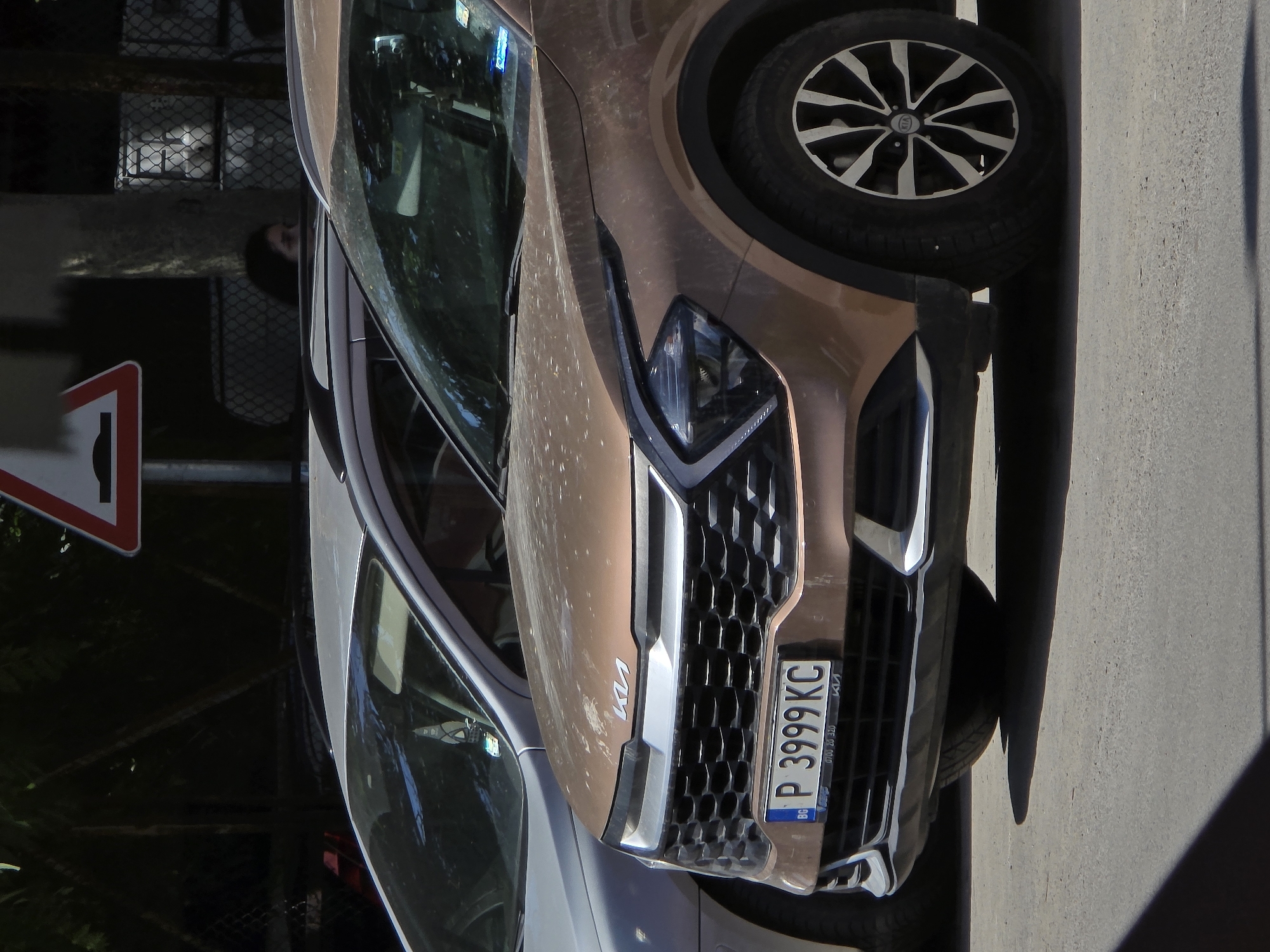 Two cars are parked on a paved surface, likely a street or parking lot, in Ruse, Bulgaria. The car in the foreground is a bronze-colored Kia Sportage, facing the viewer. It has a distinctive grille with a honeycomb pattern and sharp, angled headlights. A license plate on the front of the Kia reads "P 3999 KC". The car's hood is dusty. Behind the Kia, a silver car is visible, also parked. The background shows trees and a chain-link fence, with a red and white triangular speed bump sign mounted on a pole to the left. A person's head and shoulders are partially visible in the upper right portion of the frame, looking towards the left.