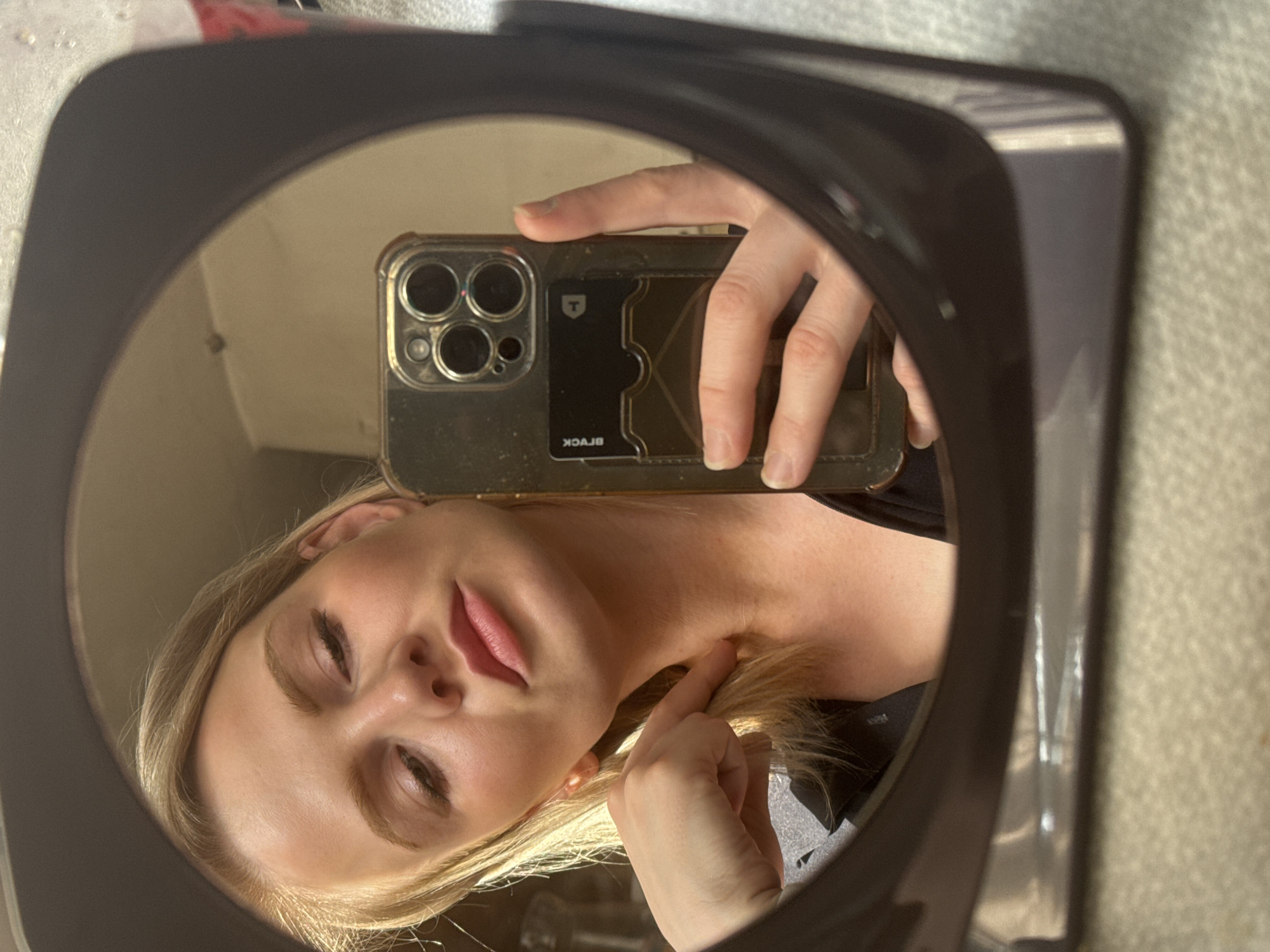 A blonde woman is taking a selfie in a black-framed mirror. She is holding a smartphone with a clear case, which appears to have a black card holder on the back containing a black card with a white "T" logo. The woman has fair skin, closed eyes, and pink lips. She is touching her neck with her right hand. The background reflected in the mirror appears to be a beige wall. The location in Saint Petersburg, Russia is not discernable from the image.