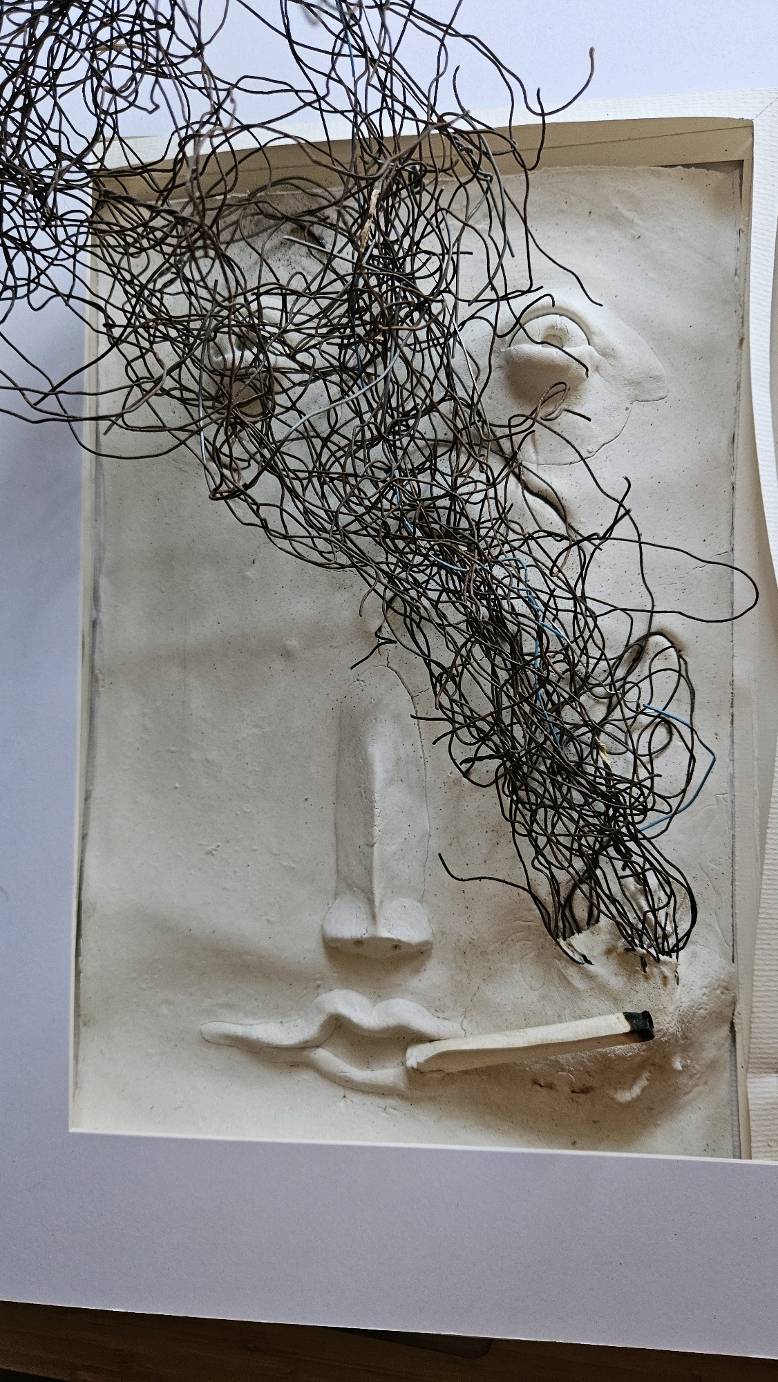 This is a close-up, eye-level, indoor photograph of an artwork. The artwork is a bas-relief sculpture on a white background, framed by a white mat and a white border. The sculpture depicts a stylized face with eyes, a nose, and lips. A cigarette is placed horizontally between the lips, with its tip in the mouth. A tangled mass of dark wires, possibly representing smoke or hair, emanates from the head area and obscures part of the face. The wires are a mix of dark metal and some blue strands. The overall color palette is monochromatic, with shades of white, grey, and black. The lighting is soft and even, highlighting the textures of the sculpture and wires. The location of this artwork in the city of Imittos, Greece, cannot be determined from the image alone.