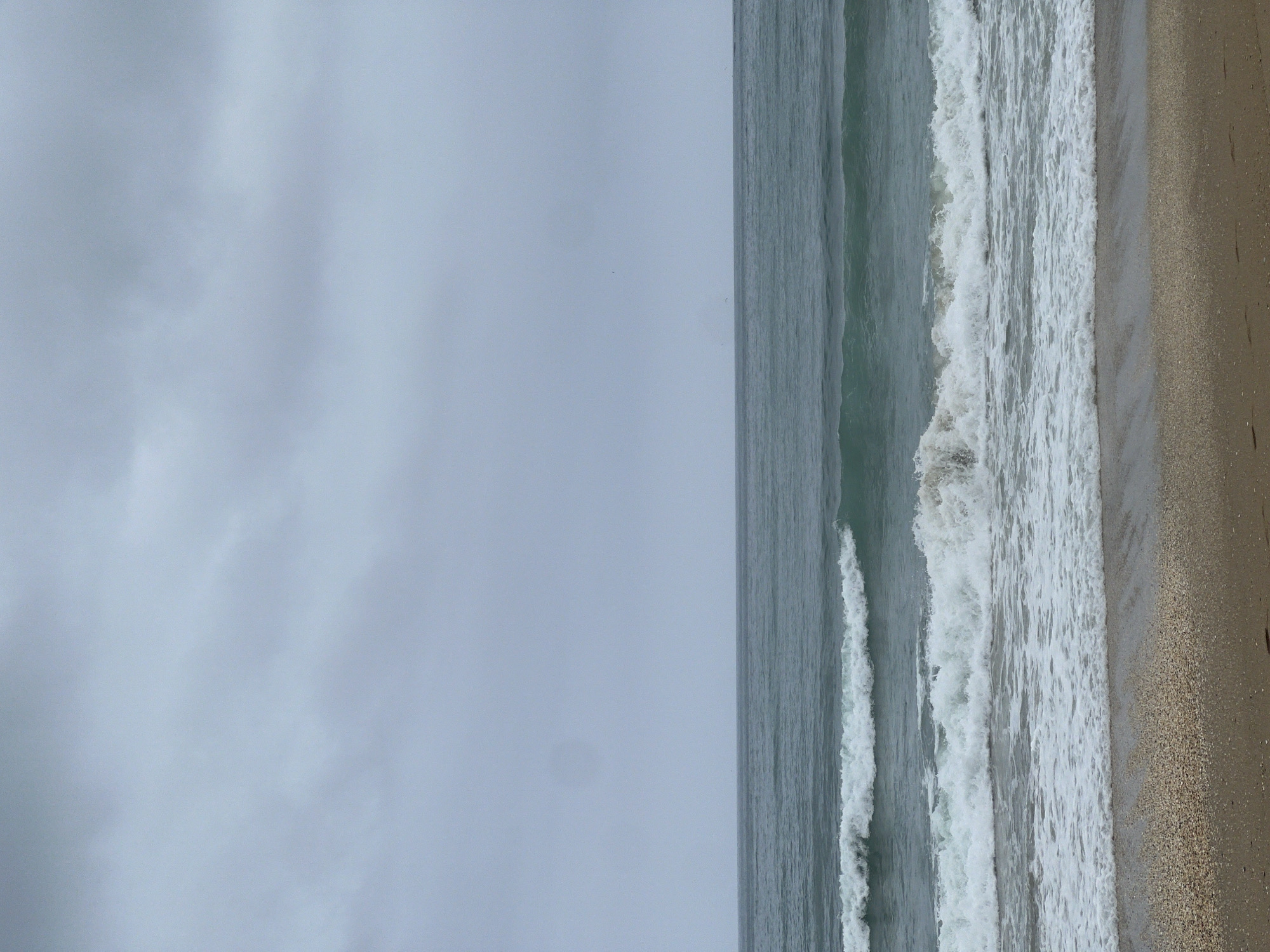The media file presents a panoramic view of a beach in M'Diq, Morocco, under an overcast sky. The image is composed of three distinct horizontal sections. The top half of the frame is dominated by a soft, uniform grey sky, indicating cloudy weather or diffused natural light.
Below the sky, the ocean stretches across the middle of the image. Further out, the water appears a deep, calm grey-blue. Closer to the shore, multiple lines of waves are visible, actively breaking and forming white, frothy foam as they roll towards the beach. The turbulent white caps contrast with the smoother water further offshore.
The bottom portion of the image displays a sandy beach, characterized by its light brown and beige tones. The sand appears textured with fine grains, suggesting a natural coastline. There are no individuals, animals, or man-made structures visible, highlighting the natural elements of the seascape. The scene conveys a sense of peaceful solitude on a cloudy day by the sea. No text is visible in the image.