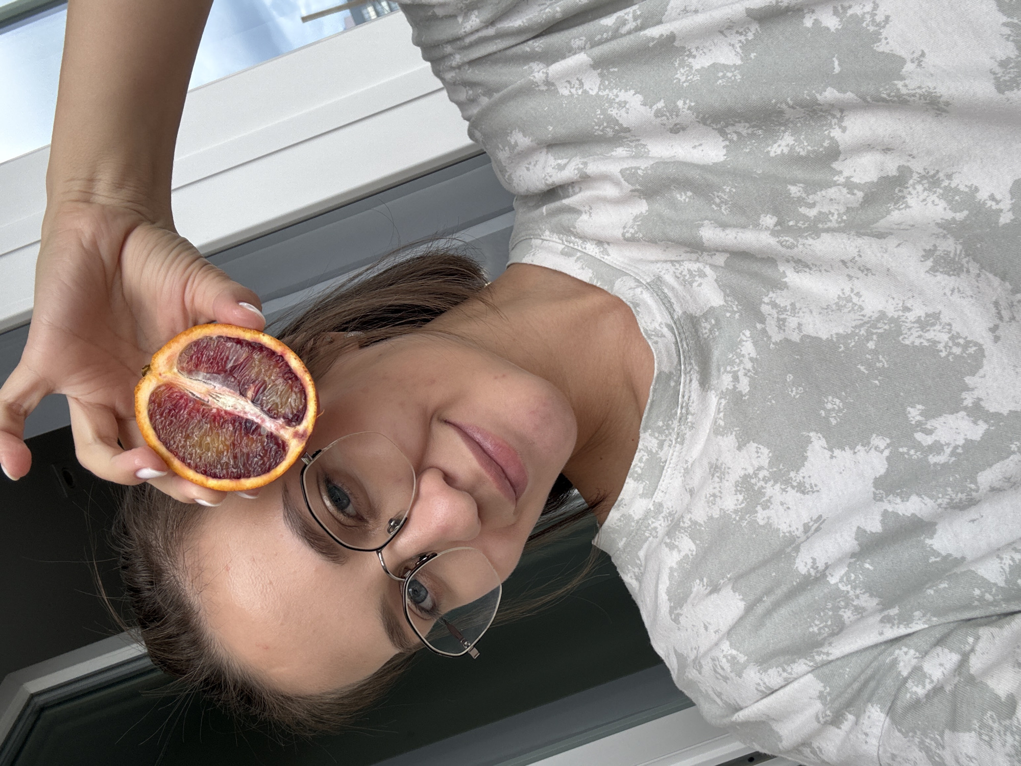 An image shows a woman holding a cut blood orange, presented with a 90-degree clockwise rotation. The woman's face is visible in the lower right portion of the frame, looking towards the viewer with a slight smile. She has brown hair, fair skin, and wears round eyeglasses with clear frames. Her eye color appears to be blue.
Her right hand holds the half blood orange near her head, showcasing its vibrant deep red and purple flesh. Her fingernails are neatly manicured with white polish. She is wearing a light-colored t-shirt with a distinctive grey and white abstract or tie-dye pattern.
The background consists of a white window frame and a view of the outdoors, revealing a blue sky with white clouds, indicating bright daylight. The scene appears to be an indoor setting next to a window. No text is visible in the image. The overall impression is casual and pleasant.
