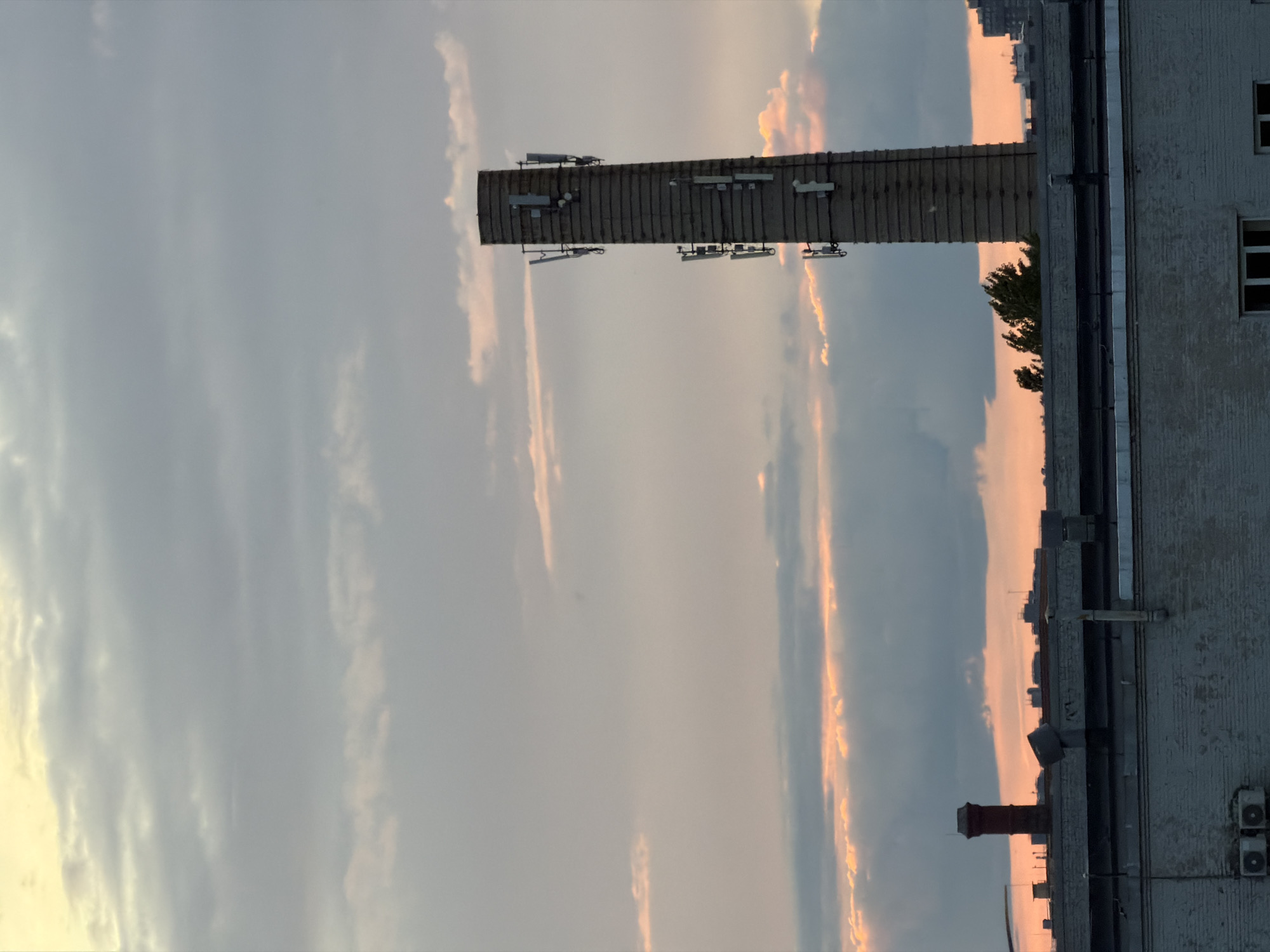 This is a vertical, eye-level, medium shot of a cityscape at dusk. In the foreground, the tops of several brick buildings are visible. The buildings appear to be industrial or commercial, with flat roofs and visible piping and ventilation units. In the midground, a tall, brick chimney stands to the right, adorned with multiple cell phone antennas. To the left of the chimney, a smaller, dark red brick chimney is visible. The background is dominated by a cloudy sky, with streaks of orange and yellow light from the setting sun filtering through. The overall impression is an urban landscape under a dramatic sky.