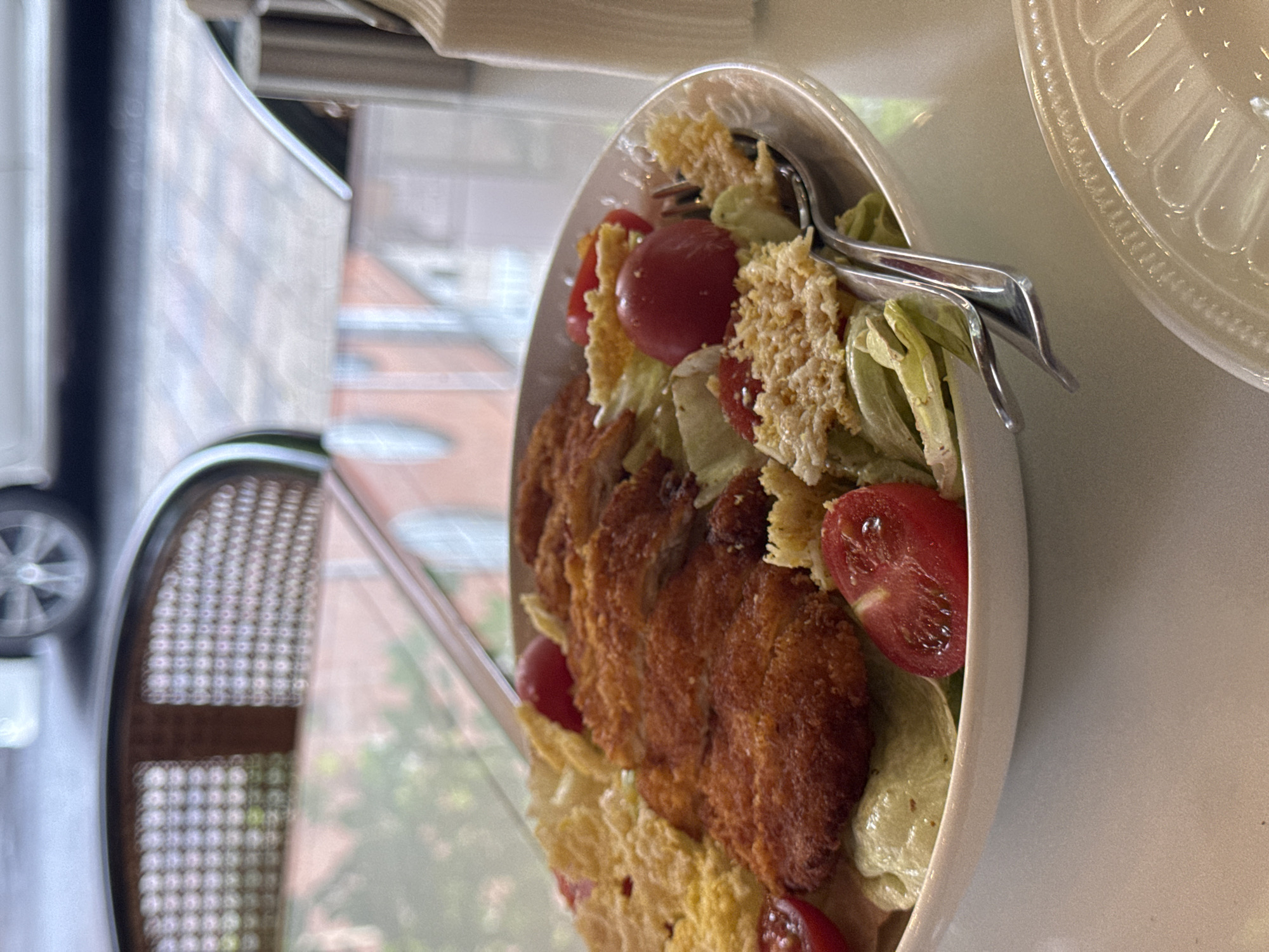 The image shows a salad in a white bowl, containing breaded meat, lettuce, cherry tomatoes, and croutons. Serving tongs rest on top of the salad. The salad is placed on a white surface near a window. Outside the window, there is a blurred view of a city street with buildings and greenery. A chair with a woven back is visible near the window. While not explicitly stated, the image appears to be taken indoors, potentially in a restaurant. It is not possible to determine the specific location in Yerevan, Armenia, based solely on this image.
