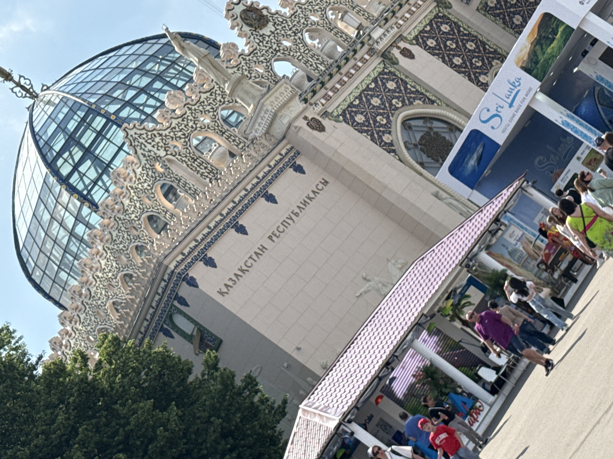 The image shows the Kazakhstan Pavilion at VDNKh (Exhibition of Achievements of National Economy) in Moscow, Russia. The pavilion features a large, geodesic dome made of glass and metal, topped by ornate decorative elements. The building's facade is adorned with intricate patterns and inscriptions in Cyrillic, which read "ҚАЗАҚСТАН РЕСПУБЛИКАСЫ" (Republic of Kazakhstan).
In the foreground, several people are visible. A person in a red t-shirt and a red cap is walking towards the left. Another person in a purple polo shirt and denim shorts is walking towards the right. Several other individuals are scattered throughout the scene, some appearing to be tourists.
In front of the pavilion, there is a shaded walkway with a purple, tiled roof. Behind this walkway, a large banner with the text "Sri Lanka" and images of the country is prominently displayed, indicating a presence or exhibition related to Sri Lanka at VDNKh. The sky is clear and blue, suggesting a sunny day. The overall impression is of an outdoor exhibition or fair, likely during the daytime.